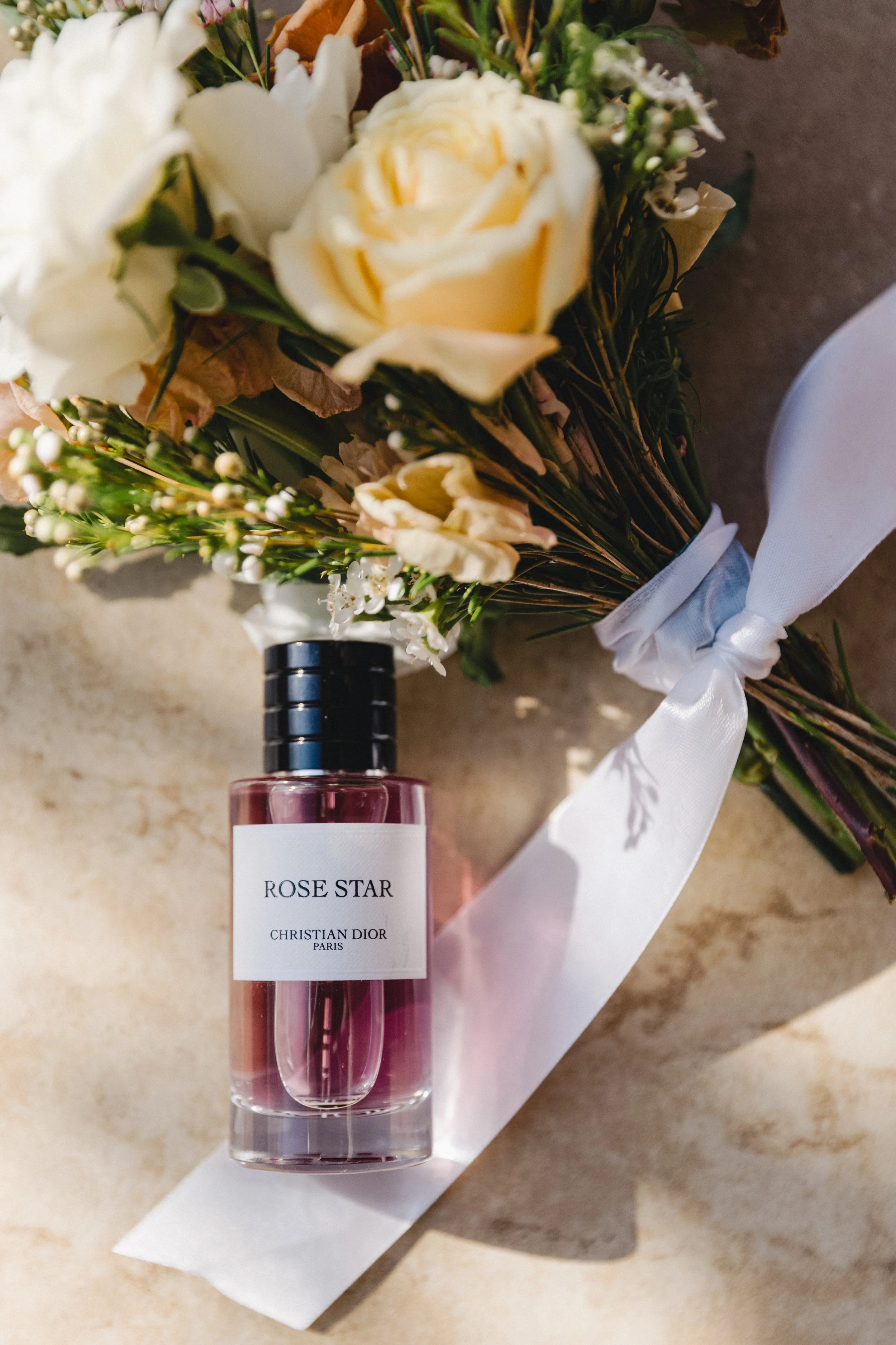 A bouquet of flowers with white and peach roses, small white filler flowers, and greenery, tied with a white ribbon, placed next to a bottle of Christian Dior Rose Star perfume on a beige surface.