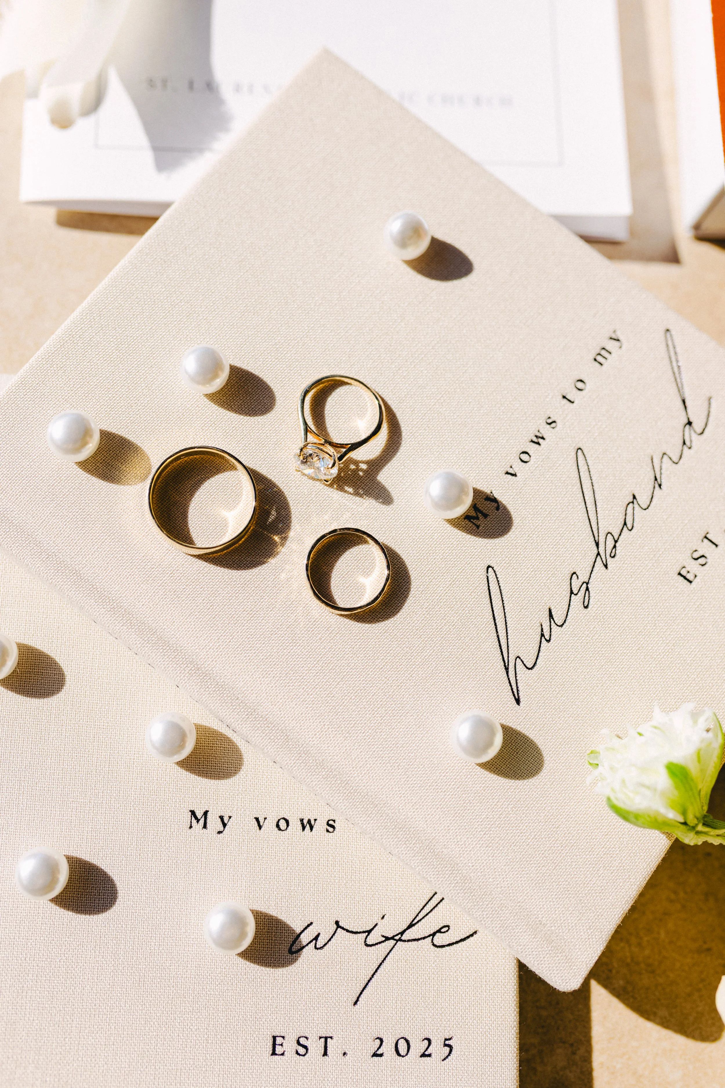 Close-up of wedding rings and pearl earrings on vow books with elegant rustic calligraphy, decorated with small pearls and a white flower.