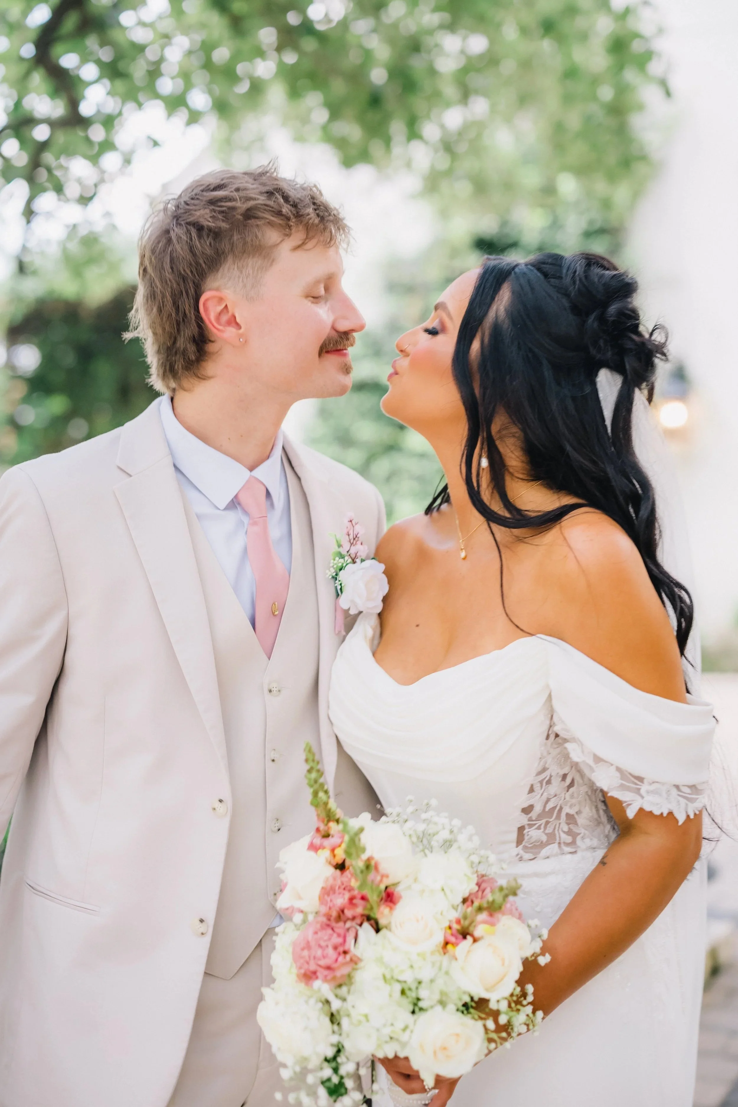 A bride and groom stand close together outdoors, with the bride holding a bouquet of white and pink flowers. The groom is wearing a light-colored suit with a pink tie, and the bride is in a white off-the-shoulder wedding dress with lace details, both leaning in for a kiss.