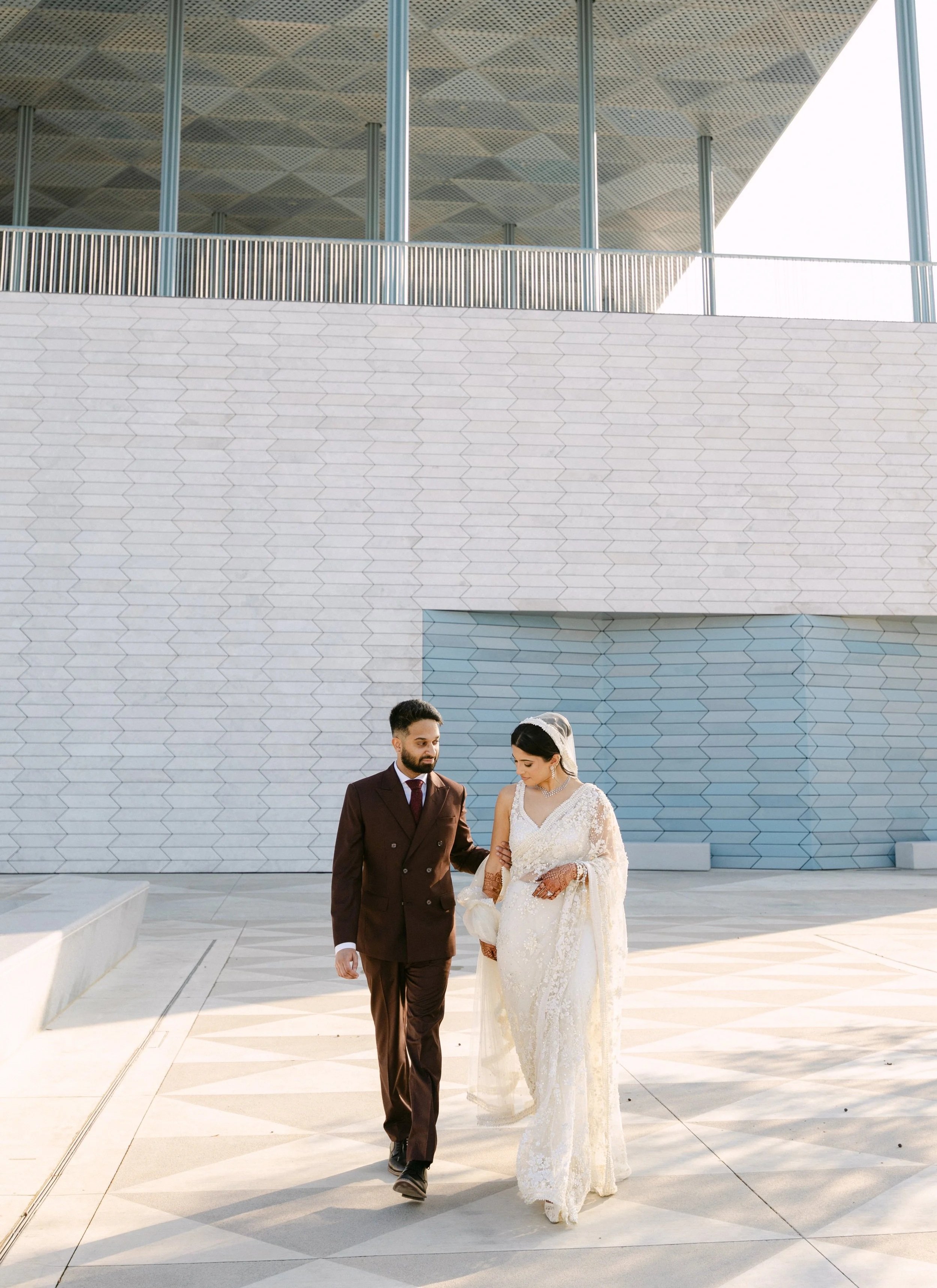 Bride and groom walking together outside in front of a modern building with hexagonal tiles on its wall. The bride is wearing a white wedding dress with lace details, and the groom is dressed in a dark brown suit.