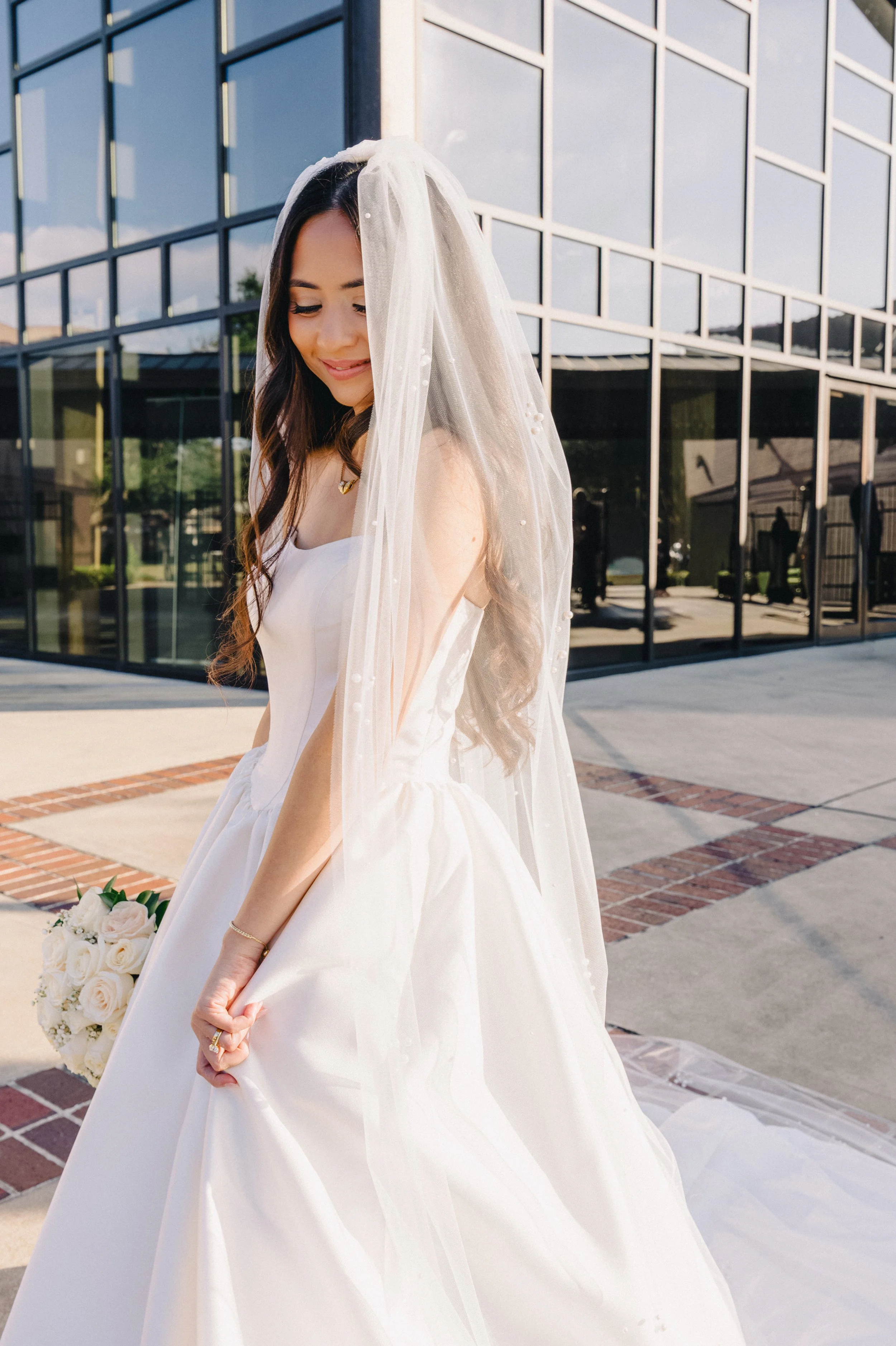A bride in a white wedding dress with a veil, holding a bouquet of white roses, smiling softly outdoors near a modern glass building.
