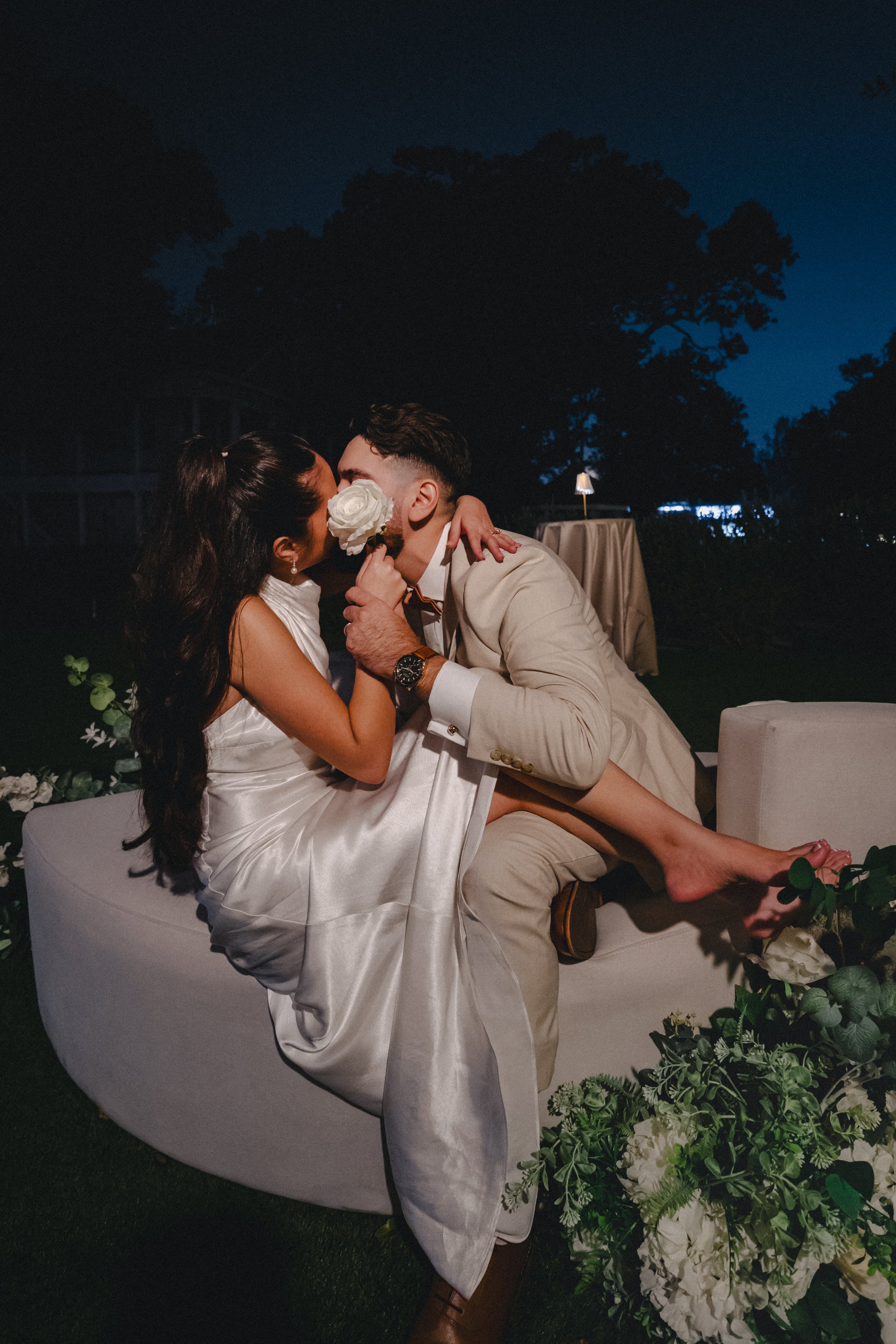 A couple sharing an intimate moment, with the woman wearing a white satin dress and the man in a beige suit, sitting on a white sofa outside at night surrounded by greenery and floral arrangements.
