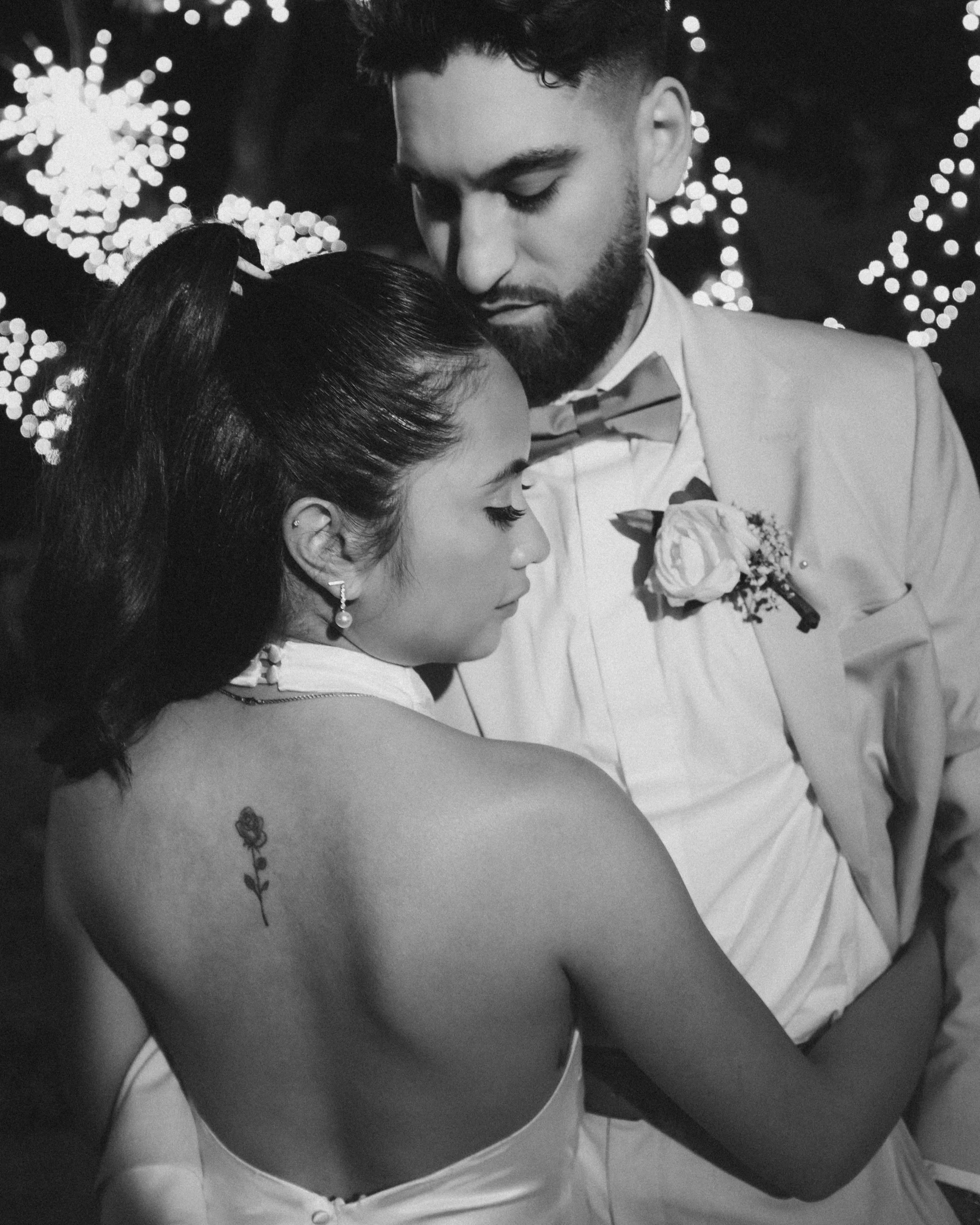 A black and white photo of a couple dressed in formal wedding attire, holding each other closely with their eyes closed, surrounded by blurred string lights.