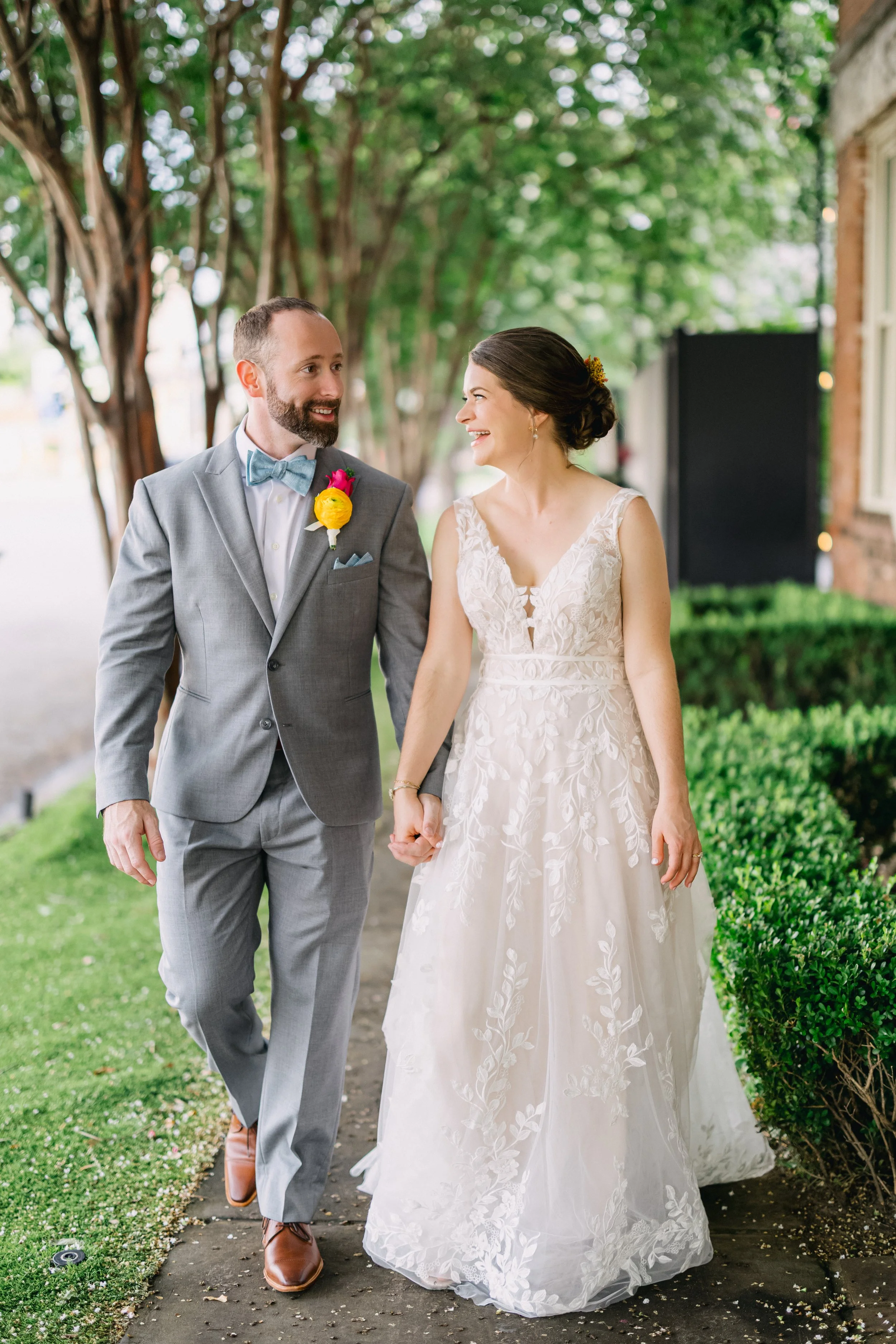 A bride and groom happily holding hands and walking outdoors, with green trees and bushes in the background.