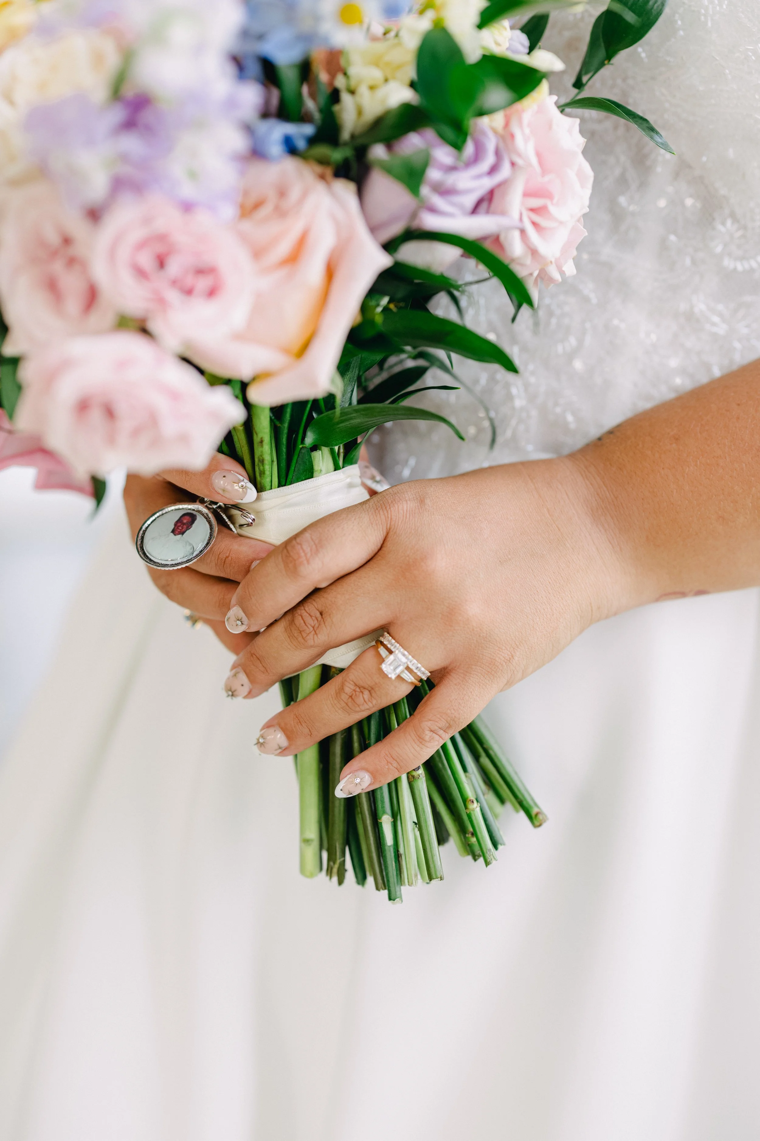 A close-up of a person's hand holding a bouquet of pink, purple, and white flowers, with a wedding ring and an oval photo ring on their fingers.