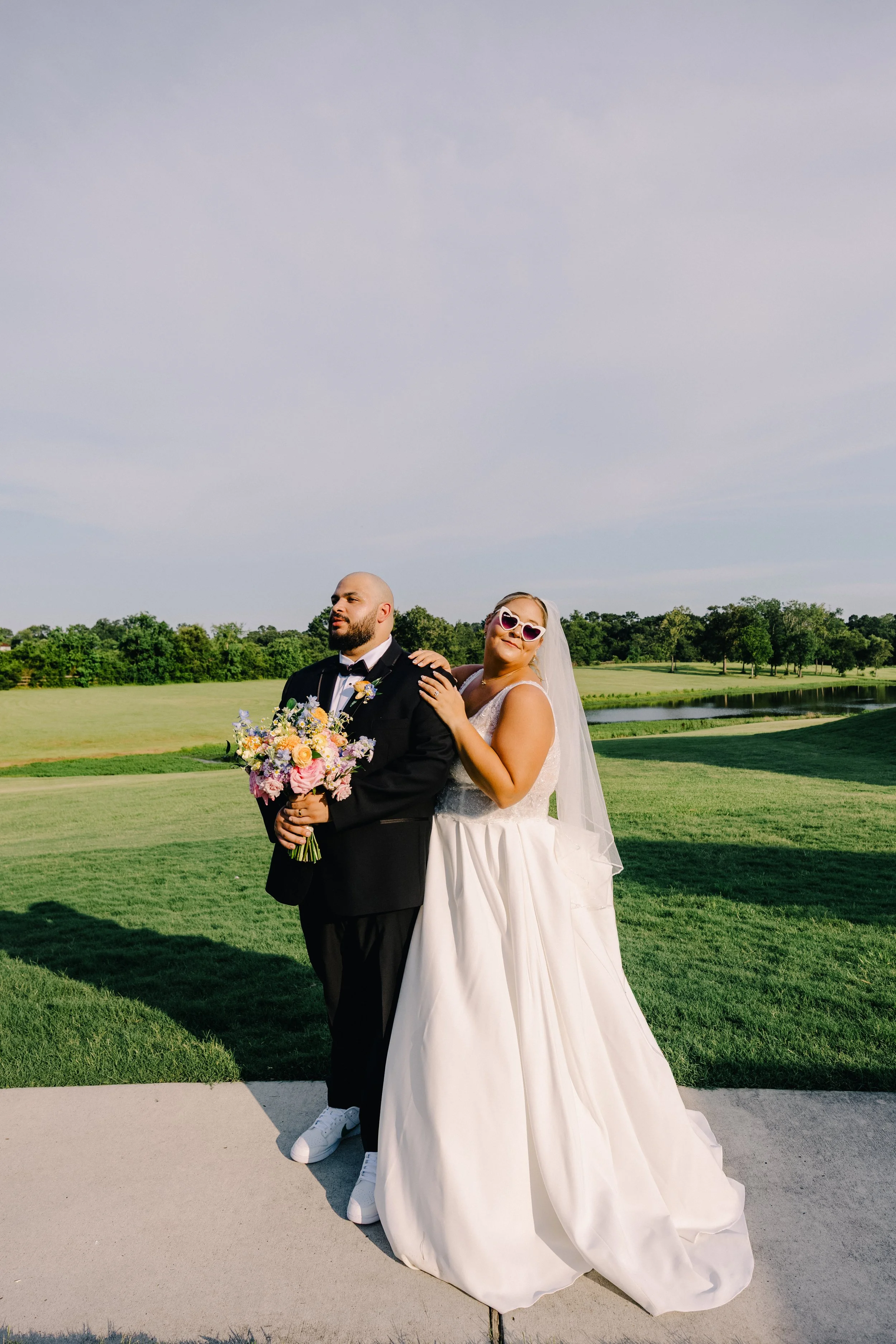 A bride and groom standing outdoors during a sunny day, with the bride wearing sunglasses and a veil, and the groom holding a bouquet of flowers.