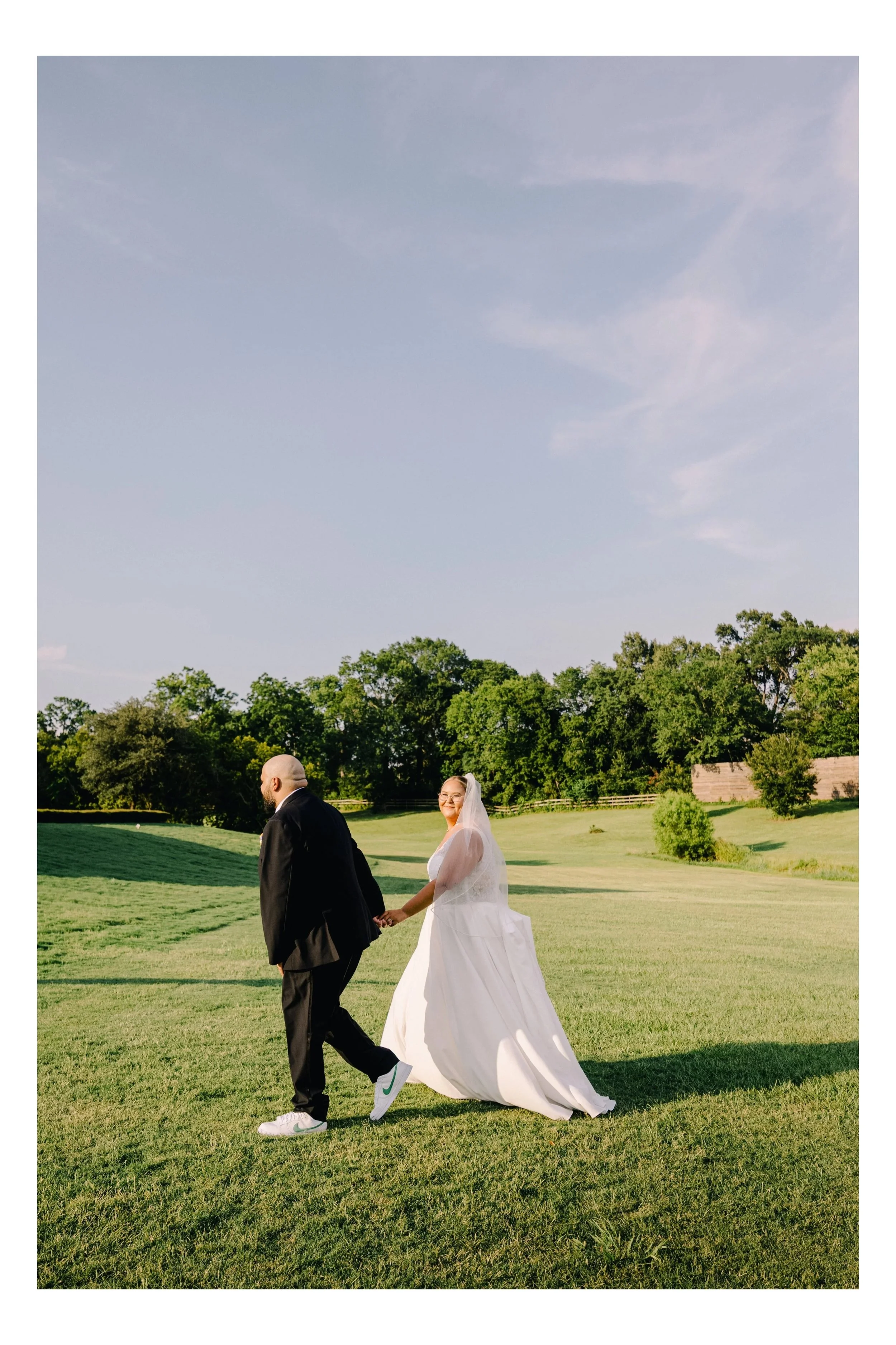 A bride and groom holding hands and walking in a lush green field during a sunny day.