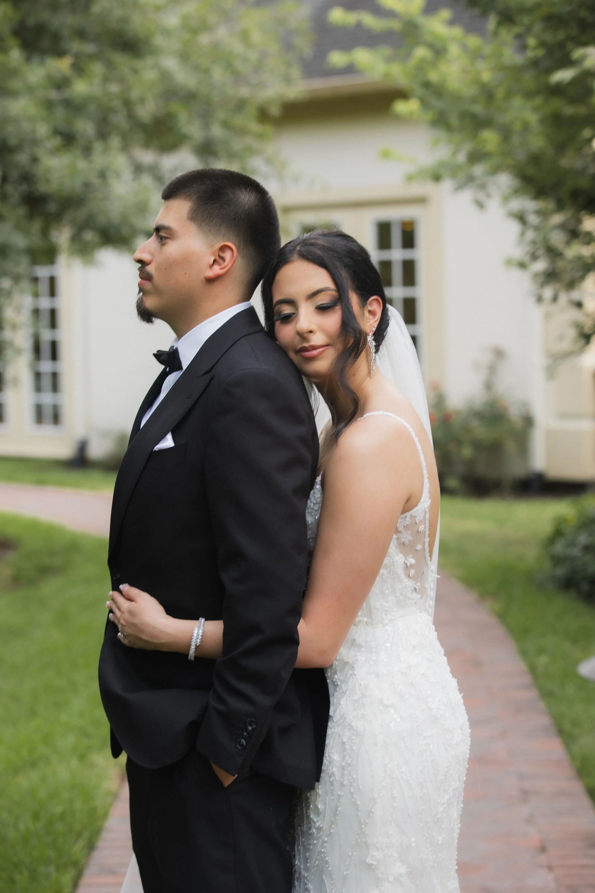 A bride and groom standing back to back outdoors, with the bride resting her head on the groom's shoulder, in a garden setting with trees and a house in the background.