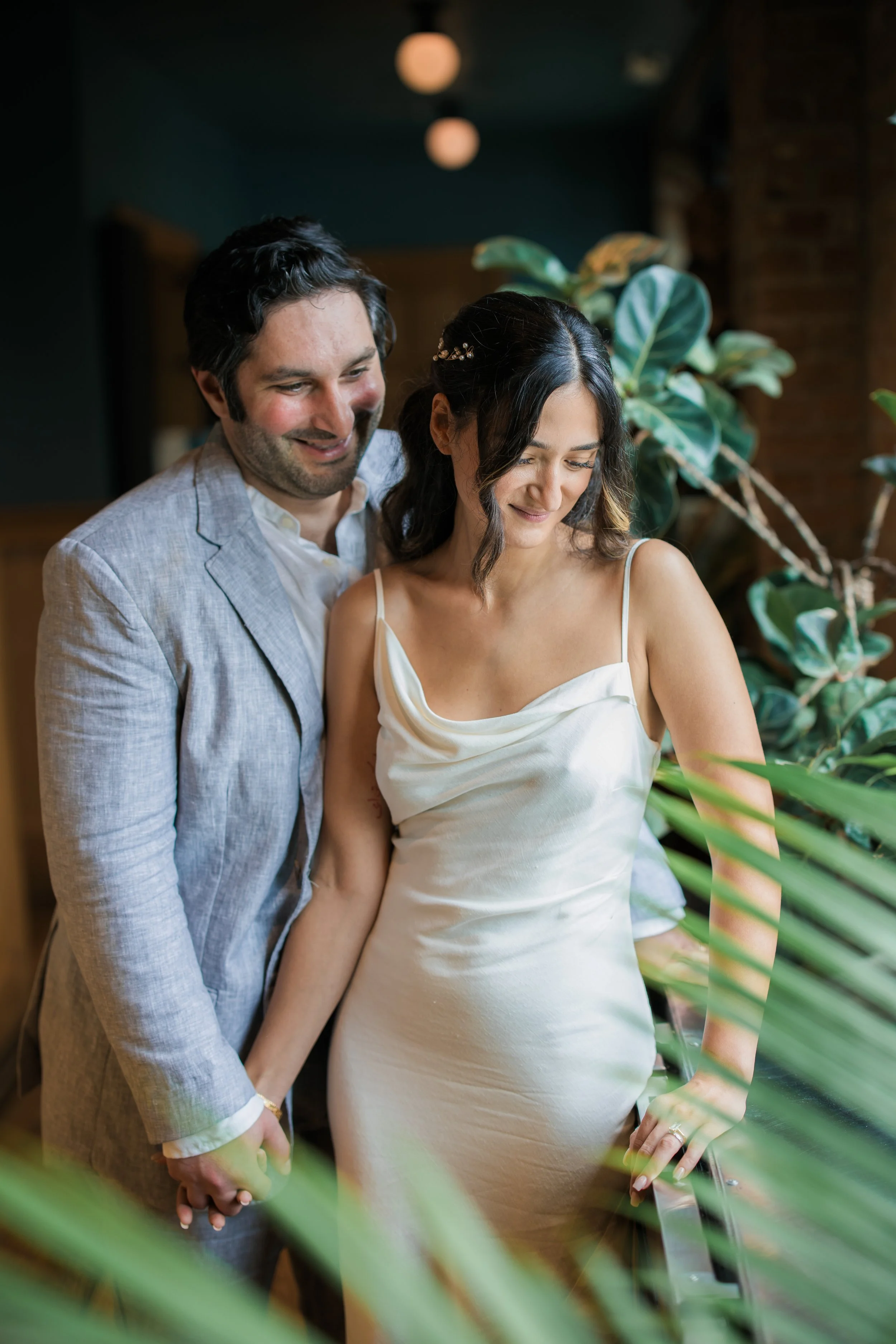 A couple is holding hands, standing close together and smiling in a cozy indoor setting with green plants and warm lighting.