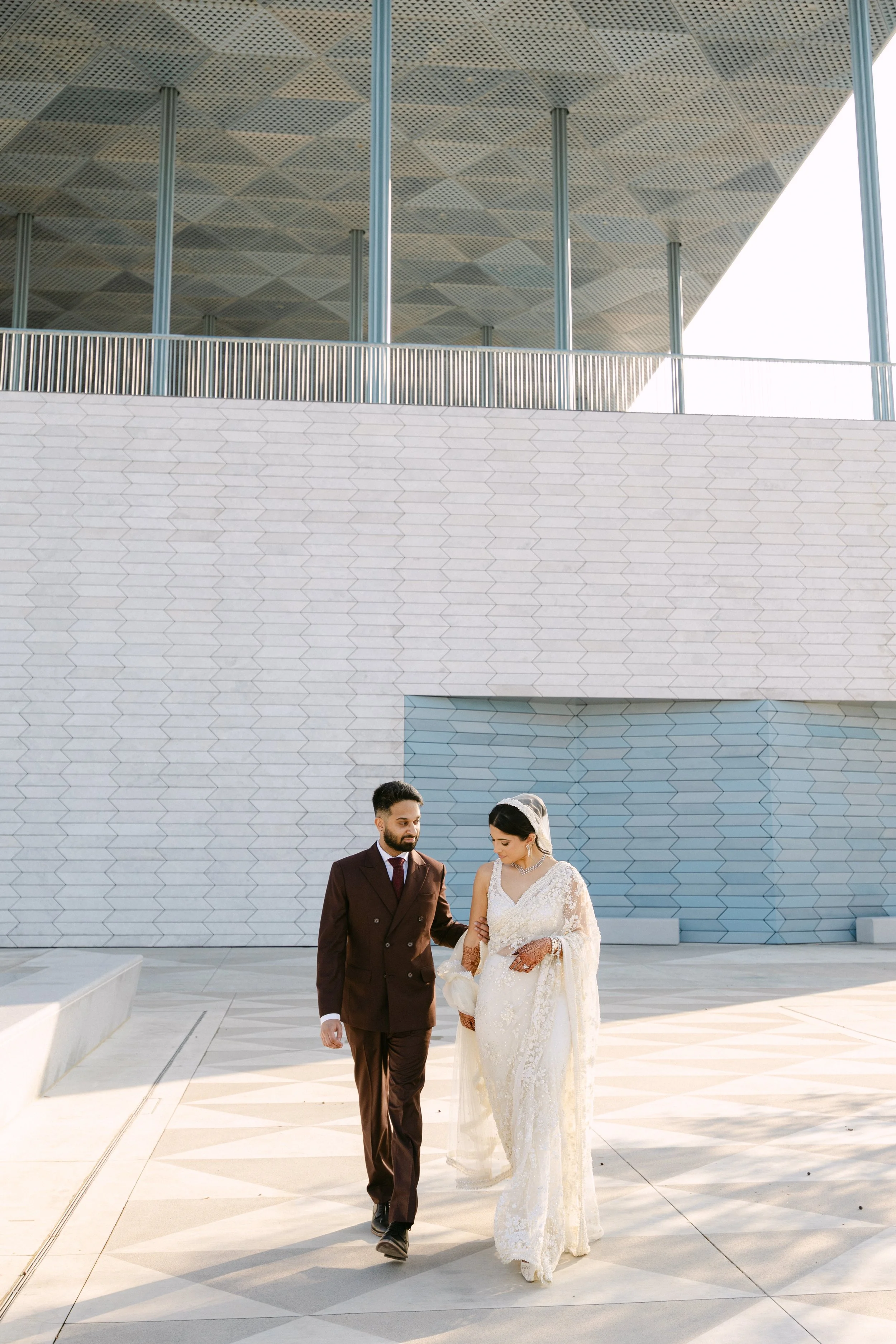 Bride and groom walking together outside in front of a modern building with hexagonal tiles on its wall. The bride is wearing a white wedding dress with lace details, and the groom is dressed in a dark brown suit.