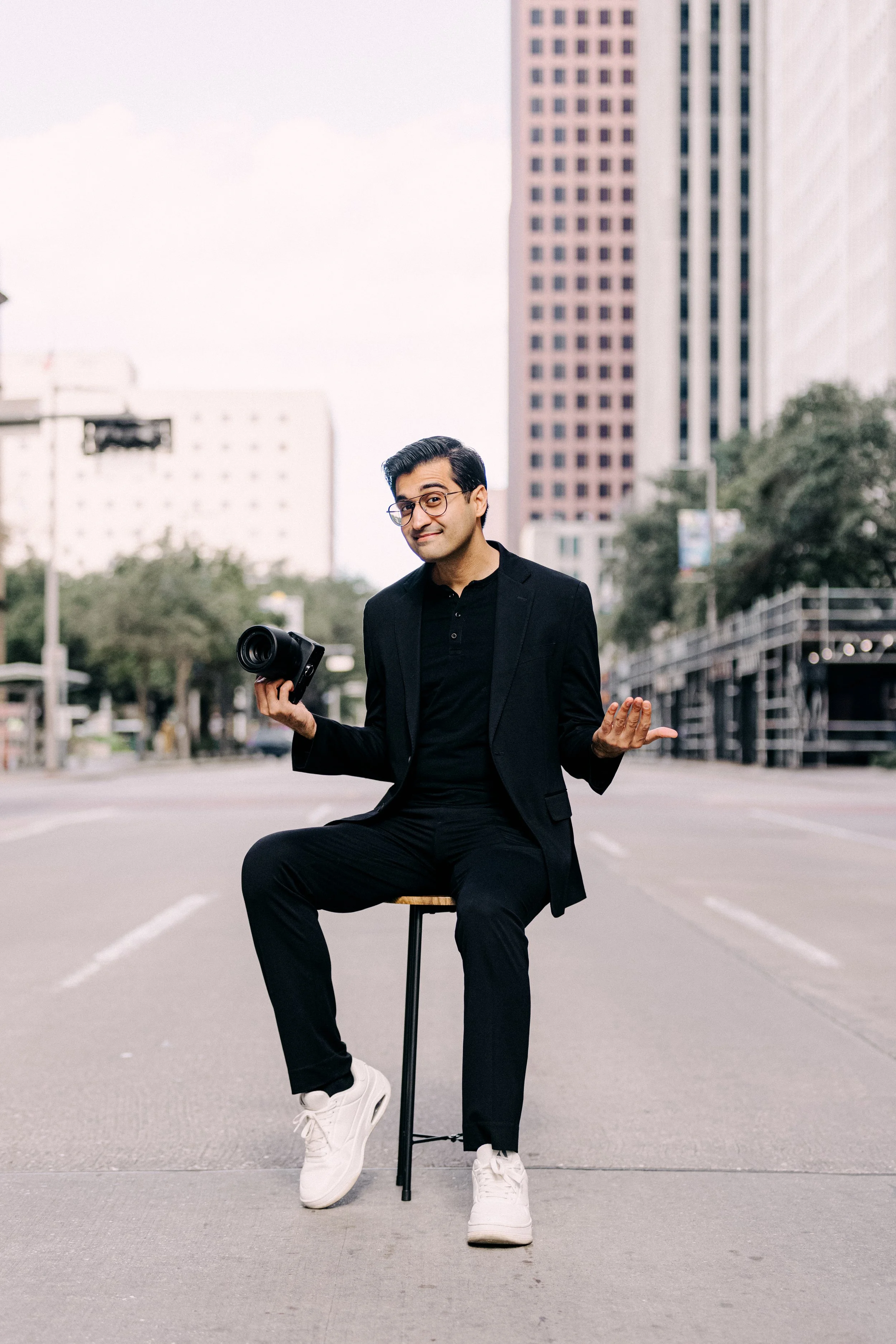 A man in a black suit and sneakers is sitting on a stool in the middle of a city street, holding a camera in one hand and gesturing with the other, with tall buildings in the background.