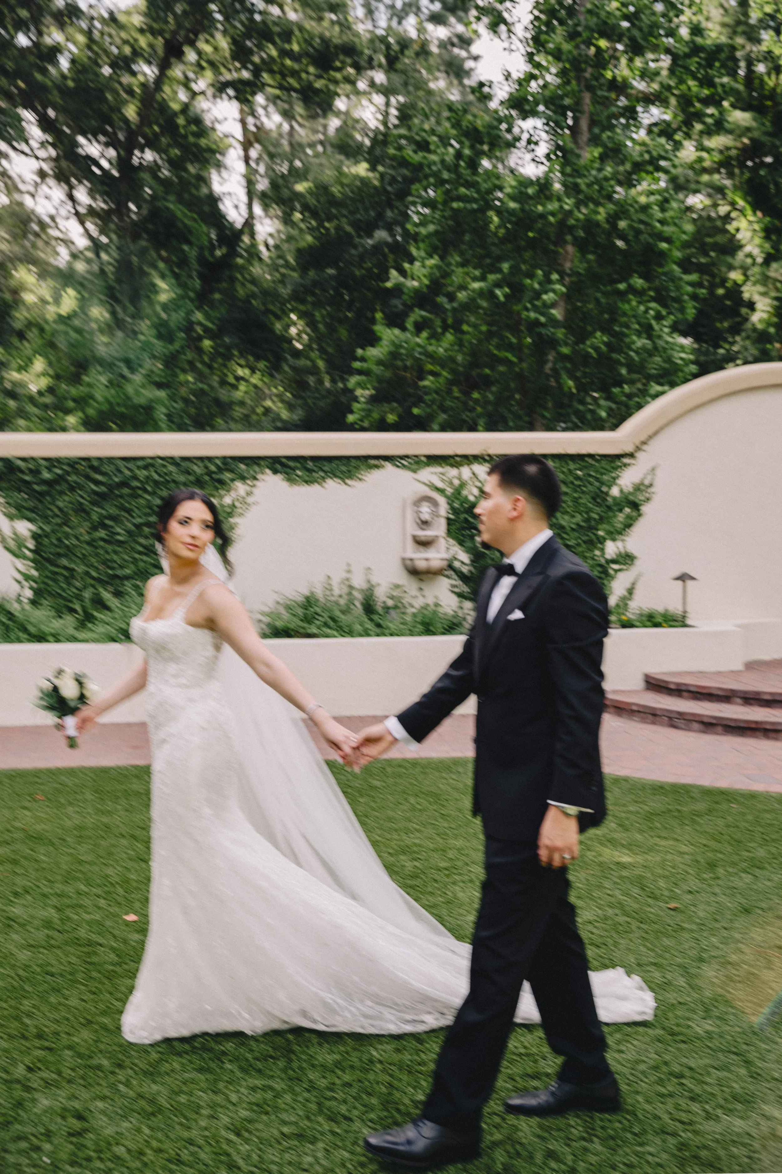 A bride and groom holding hands outdoors on a grassy area with trees and a white wall in the background.