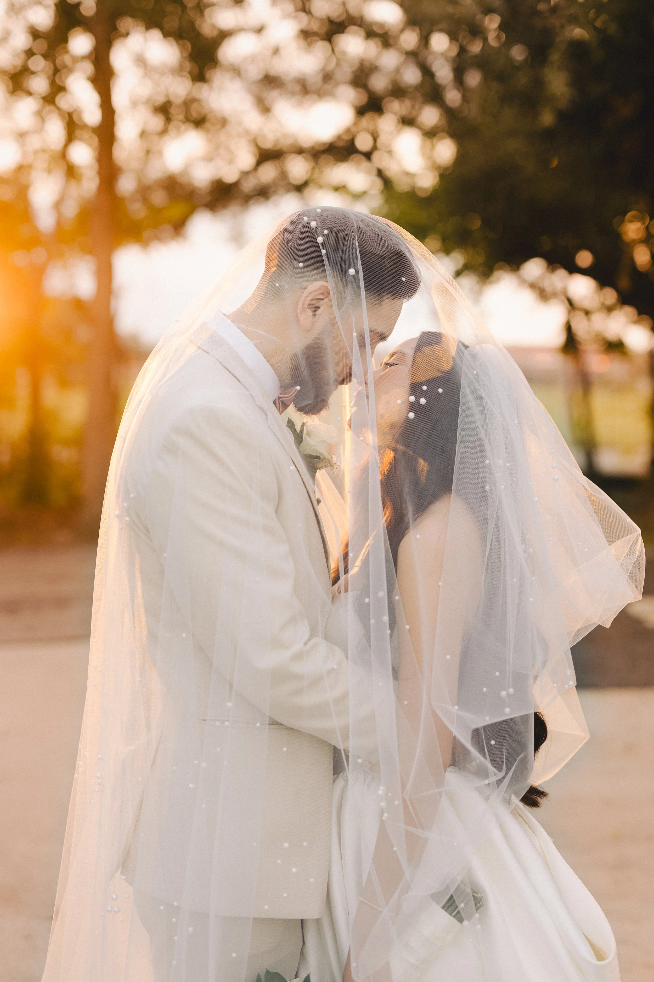 A bride and groom under a veil sharing a kiss during sunset outdoors.