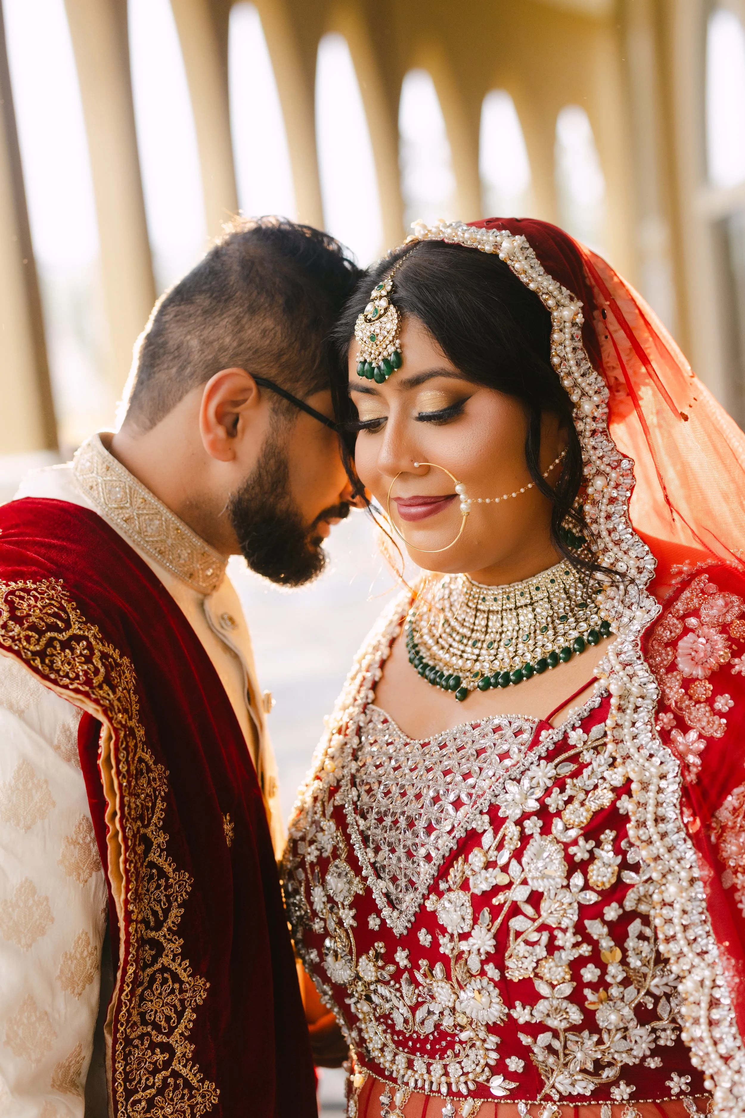 A bride and groom in traditional wedding attire, with their foreheads touching and eyes closed, standing inside with a windowed background.