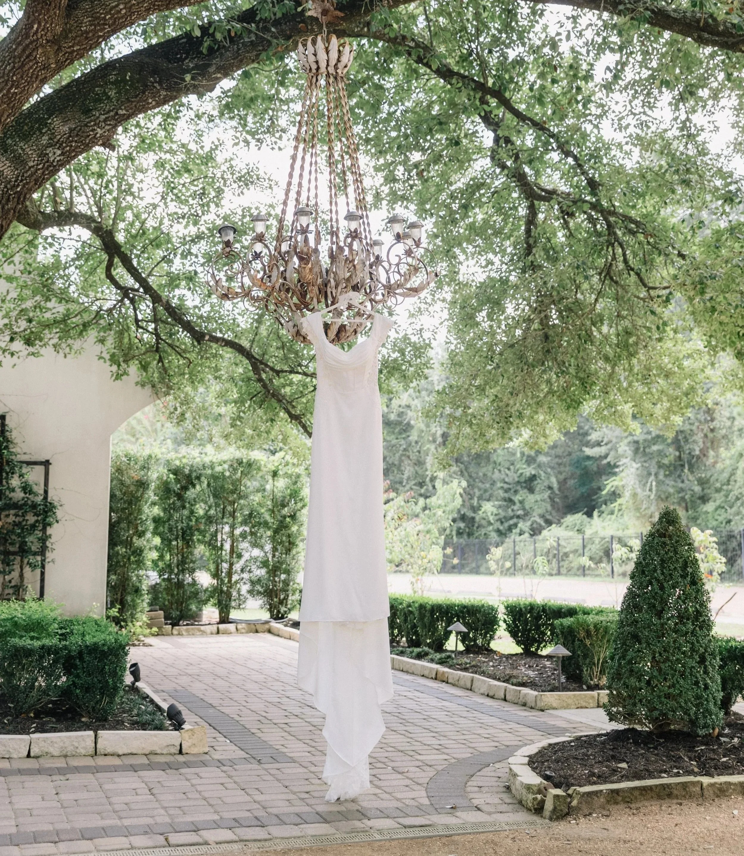 A white dress is hanging from a chandelier suspended from a tree branch outdoors.
