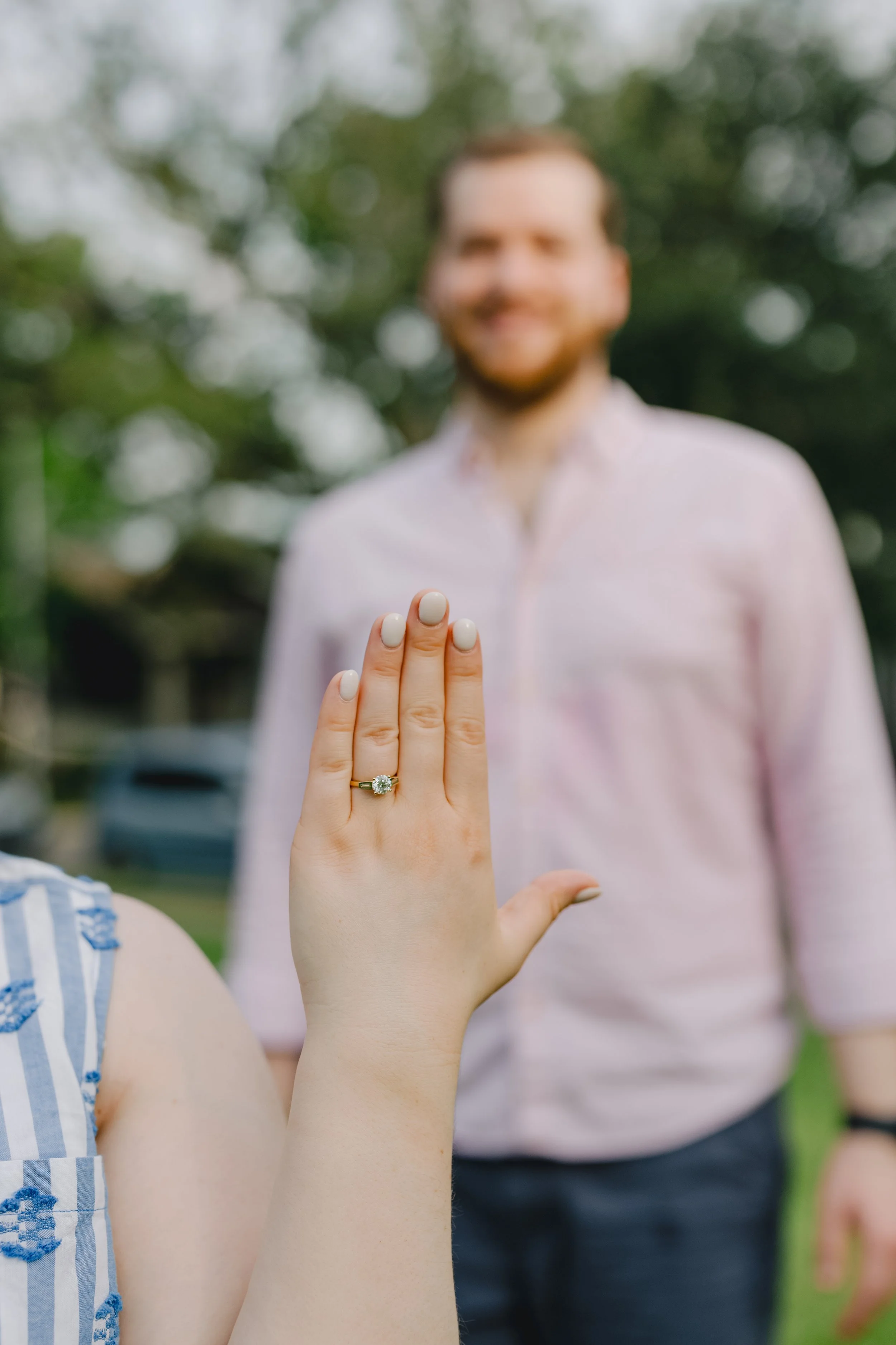 Close-up of a woman's hand displaying an engagement ring with a large central diamond and smaller side stones, with a man blurred in the background.
