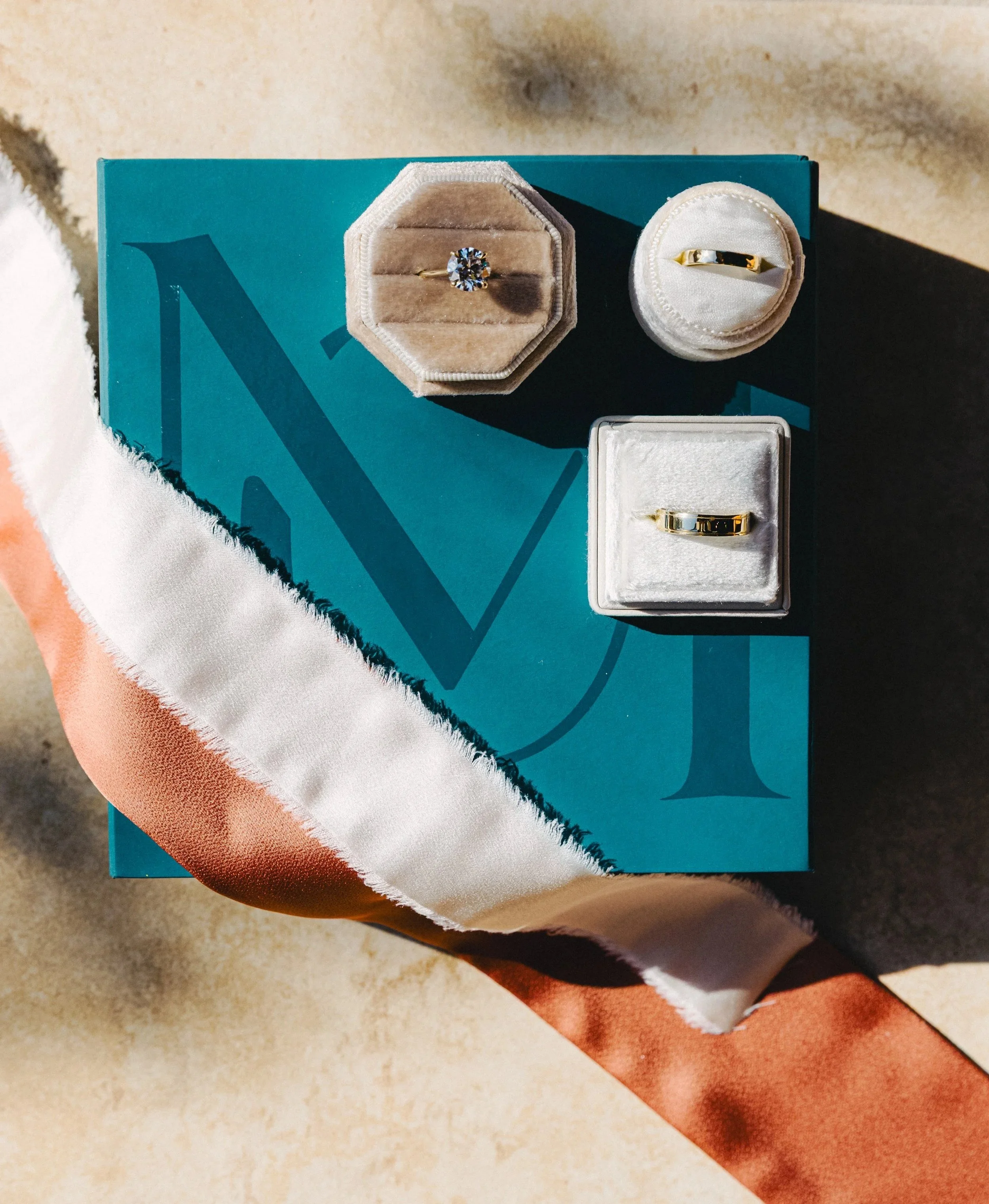 Display of three rings on a blue box with a letter 'N' and 'L' printed on it, with a satin ribbon in front of the box.