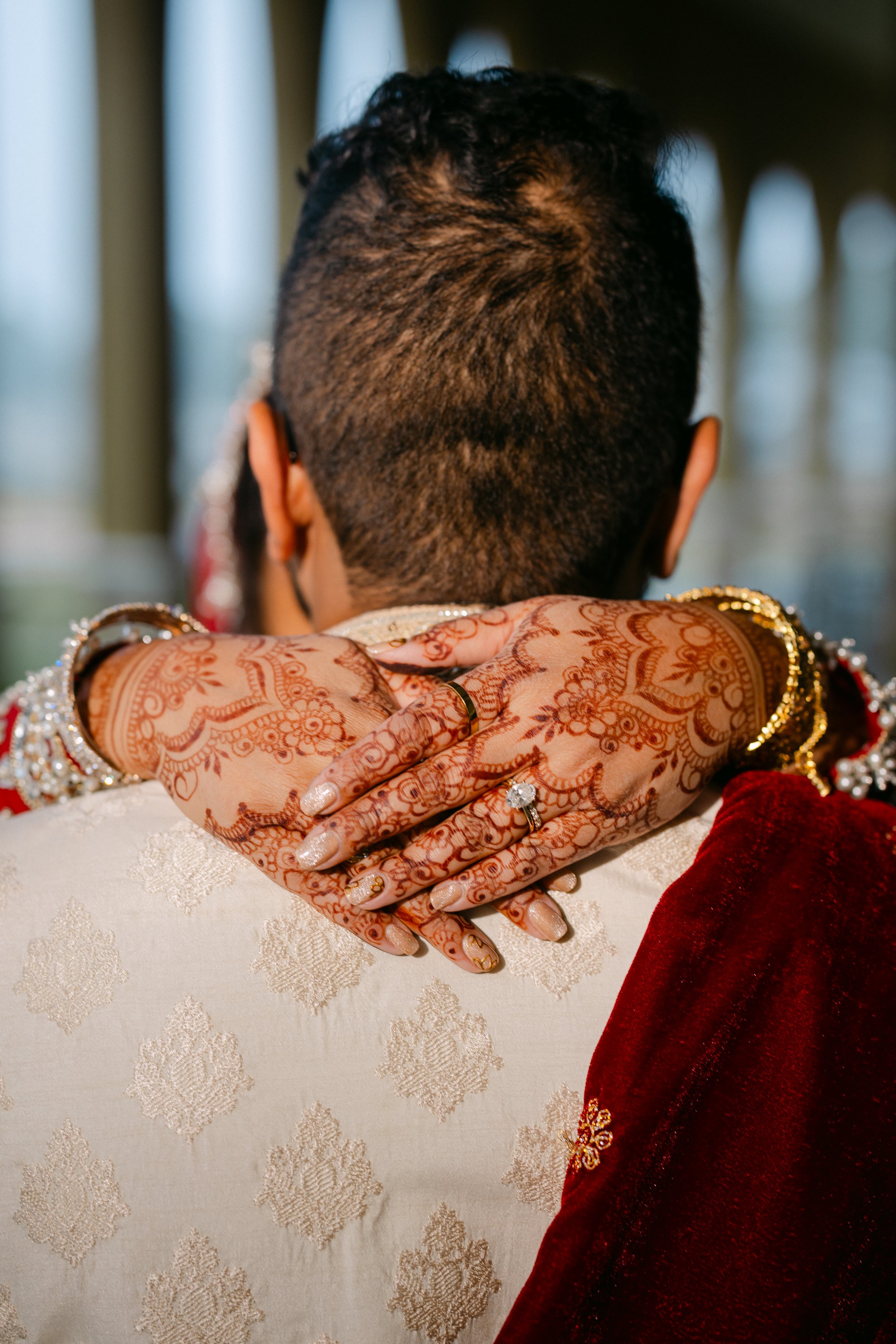 A close-up of a bride's hands resting on a groom's shoulder during a wedding, featuring intricate henna designs, jewelry, and a traditional wedding attire.