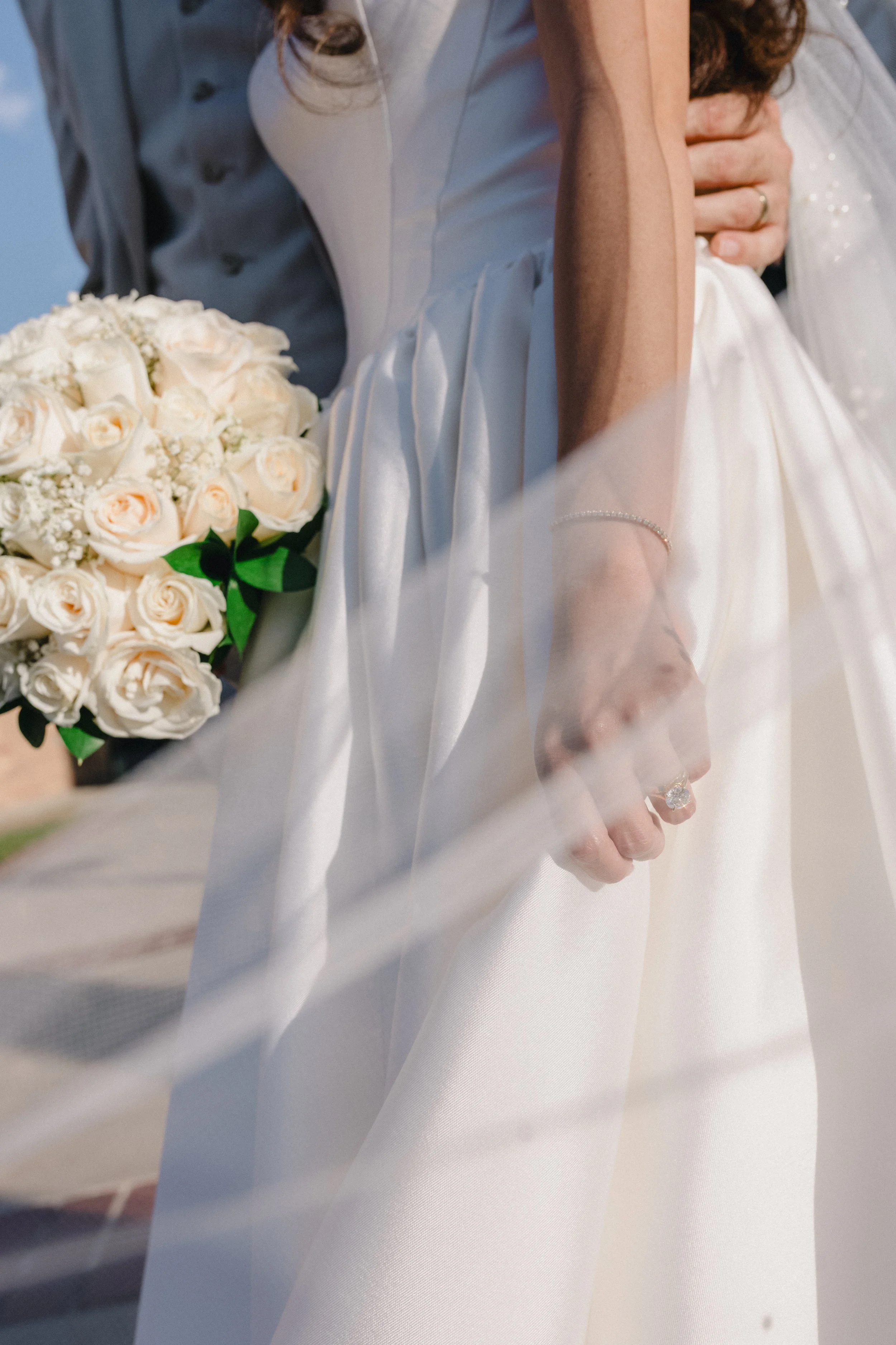 A bride holding a bouquet of white roses, wearing a white wedding dress, with a man behind her in formal attire.