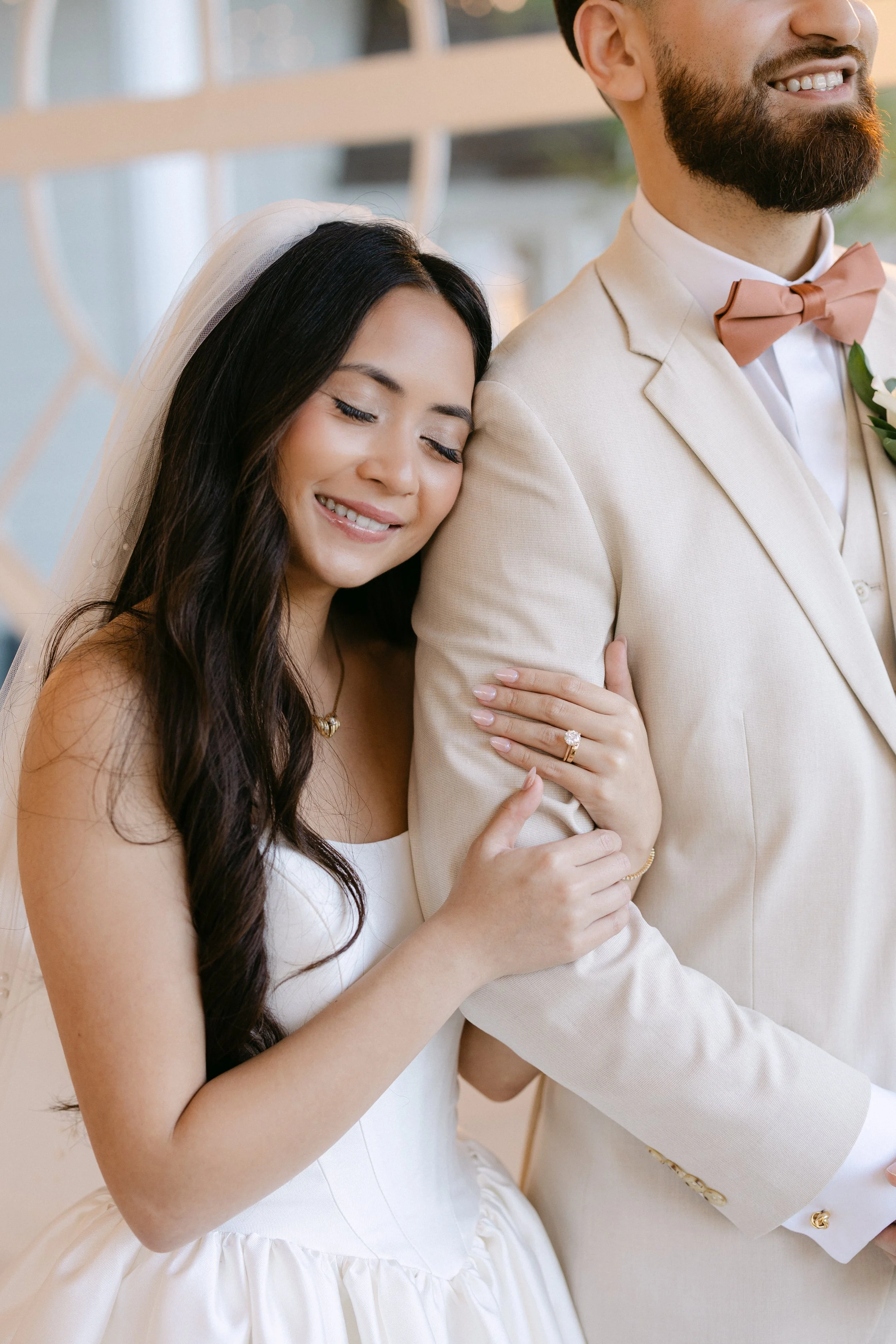 A bride with long dark hair and a white wedding dress, smiling with her eyes closed, rests her head on the arm of a groom in a cream suit with a peach bow tie, smiling slightly.