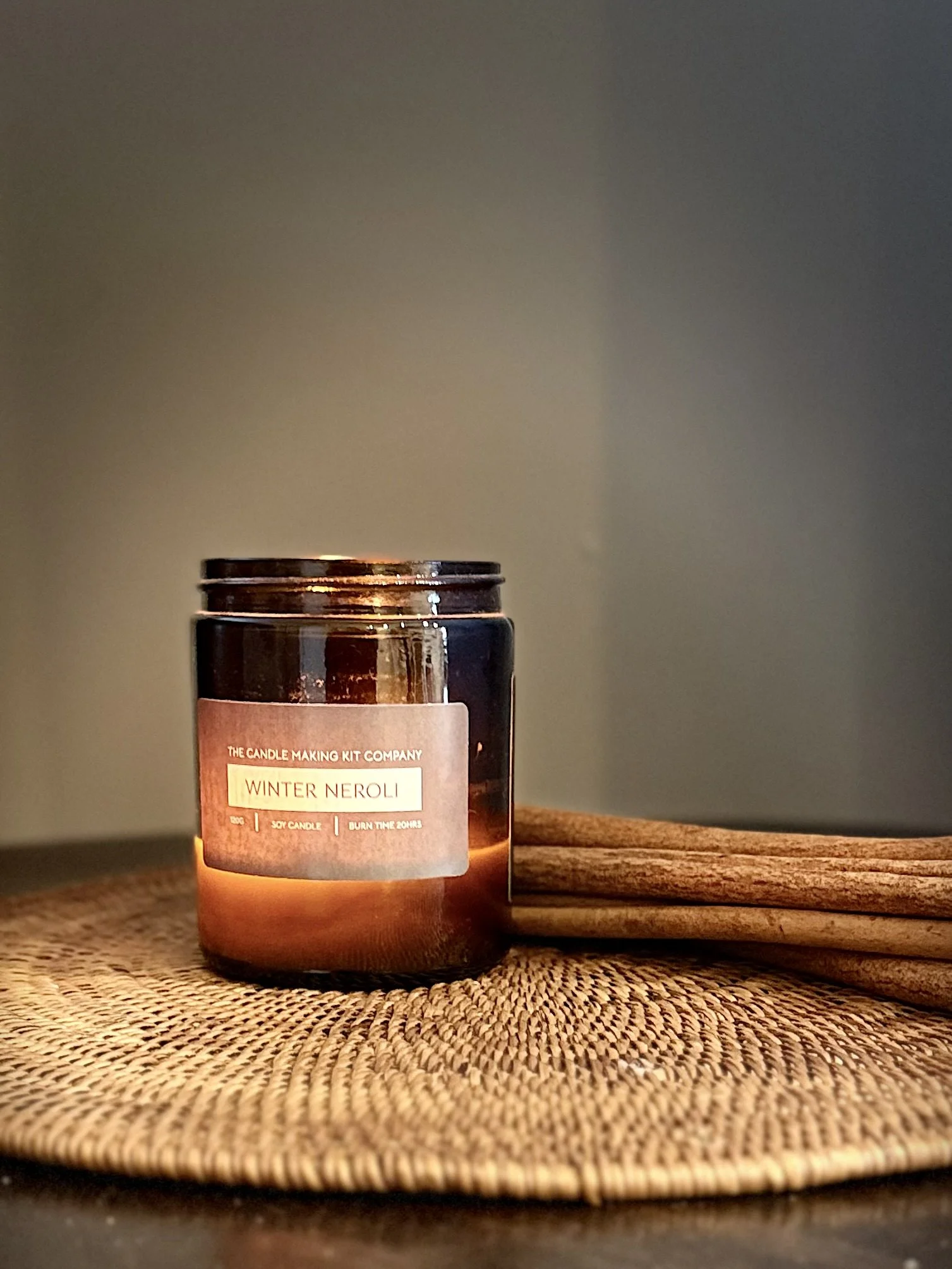 A lit candle in a brown glass jar labeled 'Winter Neroli' on a woven mat with a wooden spoon or spatula beside it, against a plain wall.