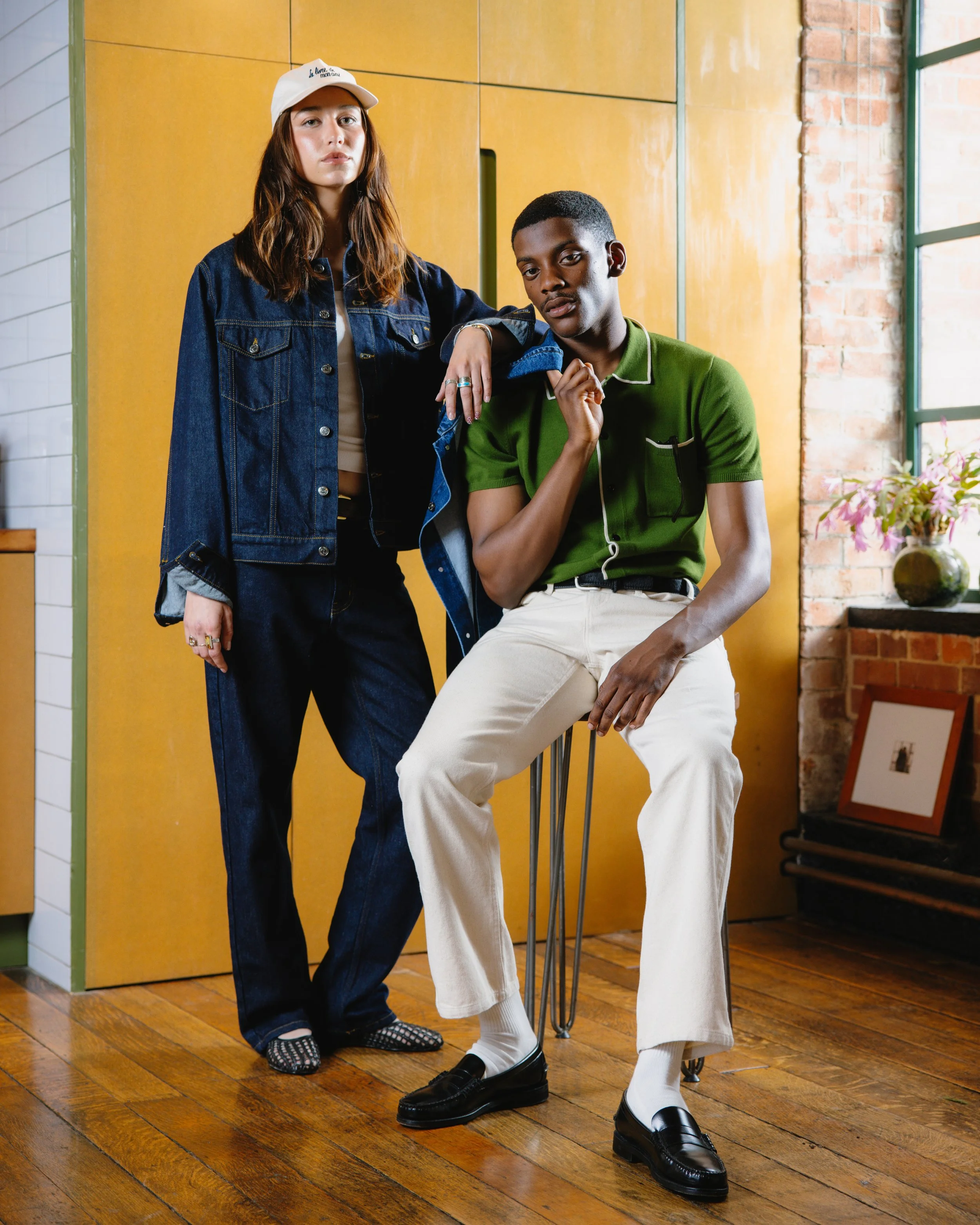 Two young adults posing indoors, one standing with hand on shoulder of the other who is sitting on a stool. The woman is wearing a white cap, denim jacket, and jeans, while the man is wearing a green polo shirt, cream pants, and black loafers.