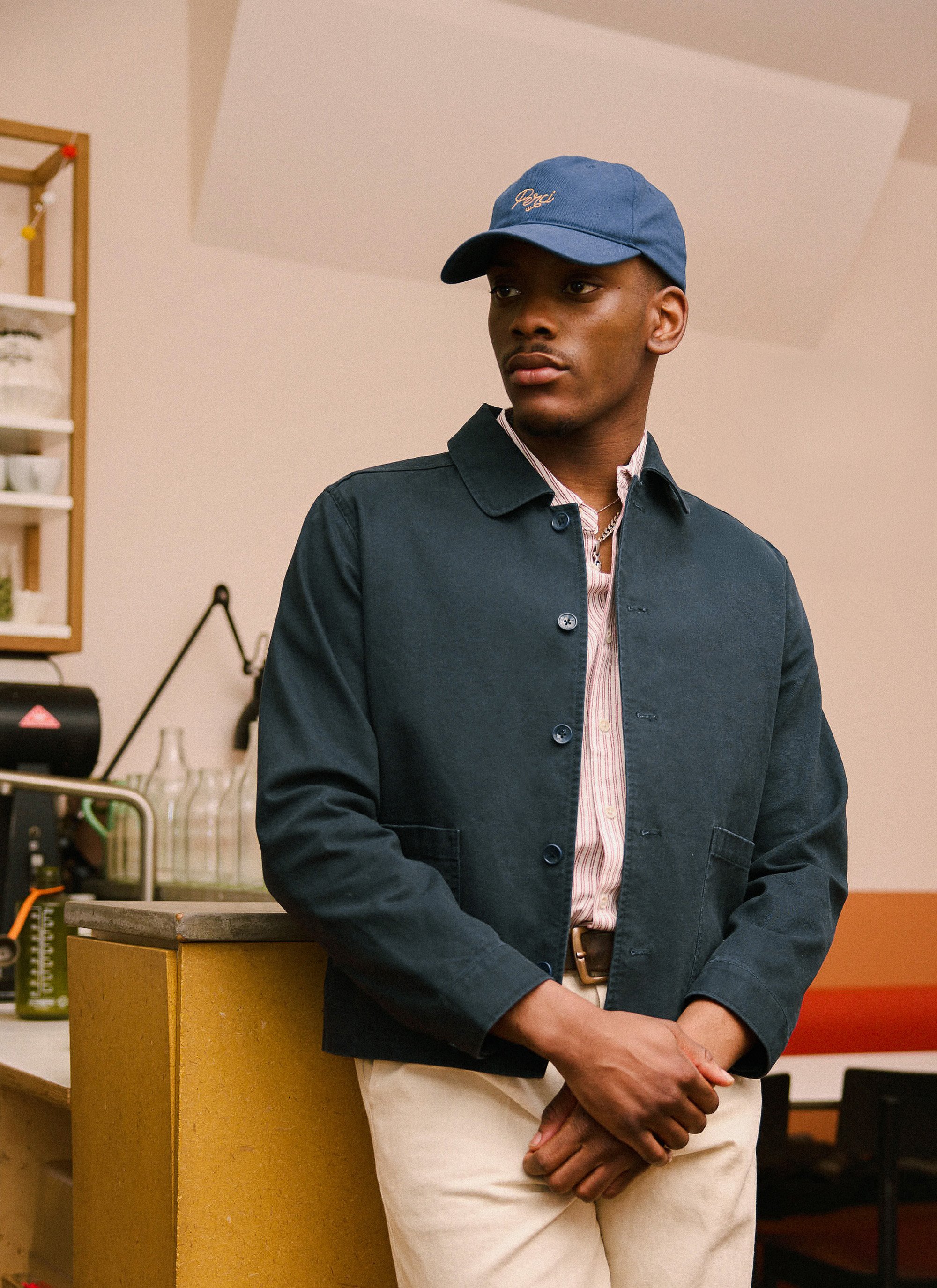 Young man in blue baseball cap, navy blazer, and beige pants standing in a room with kitchen items and shelves in the background.