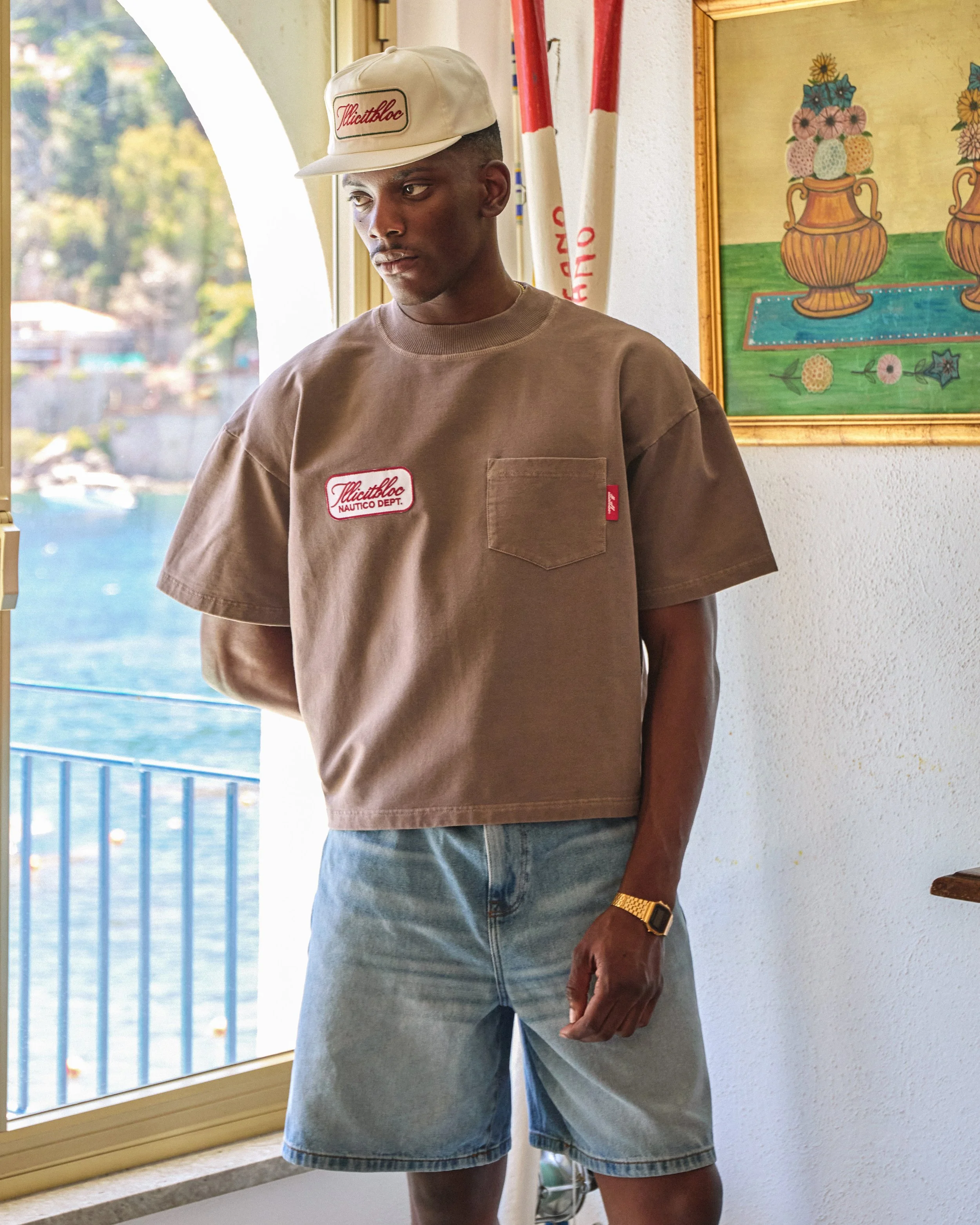 Young man standing indoors near a window, wearing a beige T-shirt with a pocket and patches, denim shorts, a beige cap with red embroidery, and a gold watch, with an outdoor water scene and colorful paintings in the background.