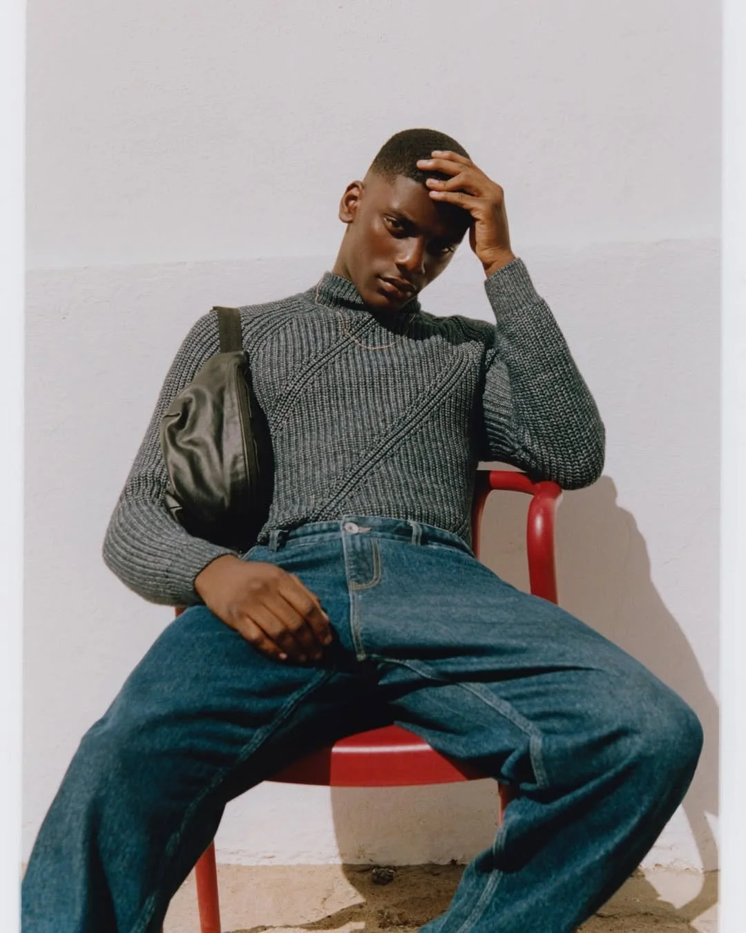 A young man sitting on a red chair against a plain white wall, wearing a gray patterned sweater and blue jeans, with a black backpack slung over his shoulder. He is leaning back with one hand resting on his forehead and the other on his lap, striking