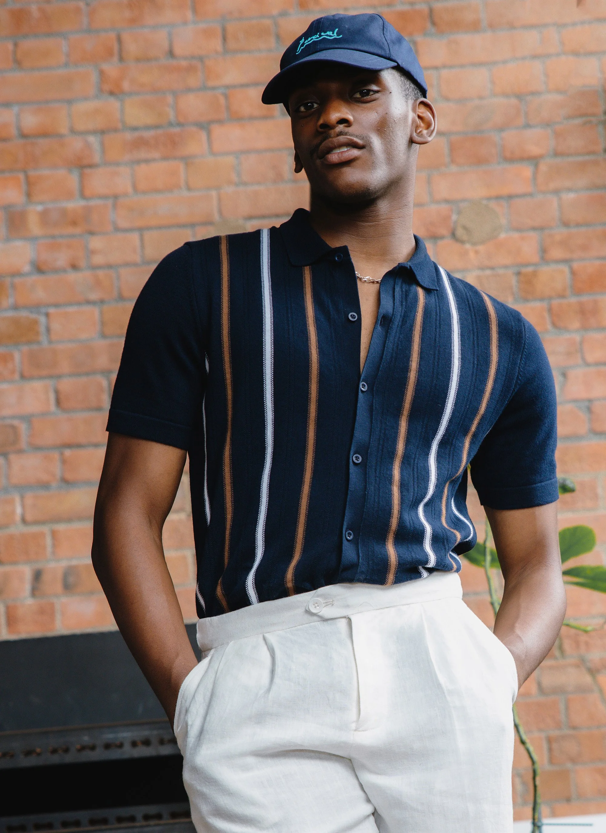 Young man wearing a navy baseball cap, navy striped short-sleeve button-up shirt, and light-colored pants, standing against a brick wall with hand in pocket.