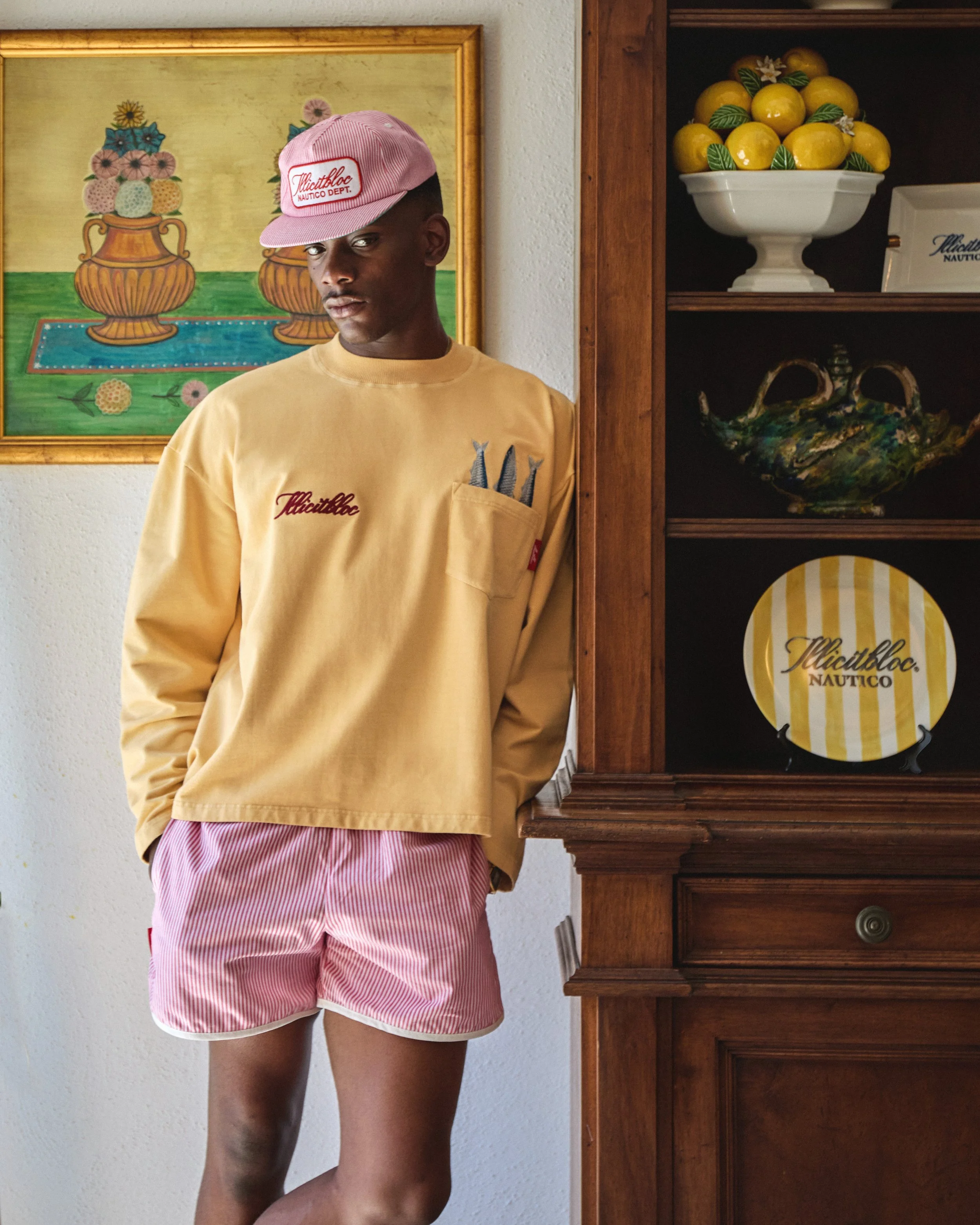 Young man wearing a pink baseball cap, yellow sweatshirt with a pocket holding pens, and pink striped shorts, standing beside a wooden cabinet with decorative plates and a bowl of lemons in a home interior.