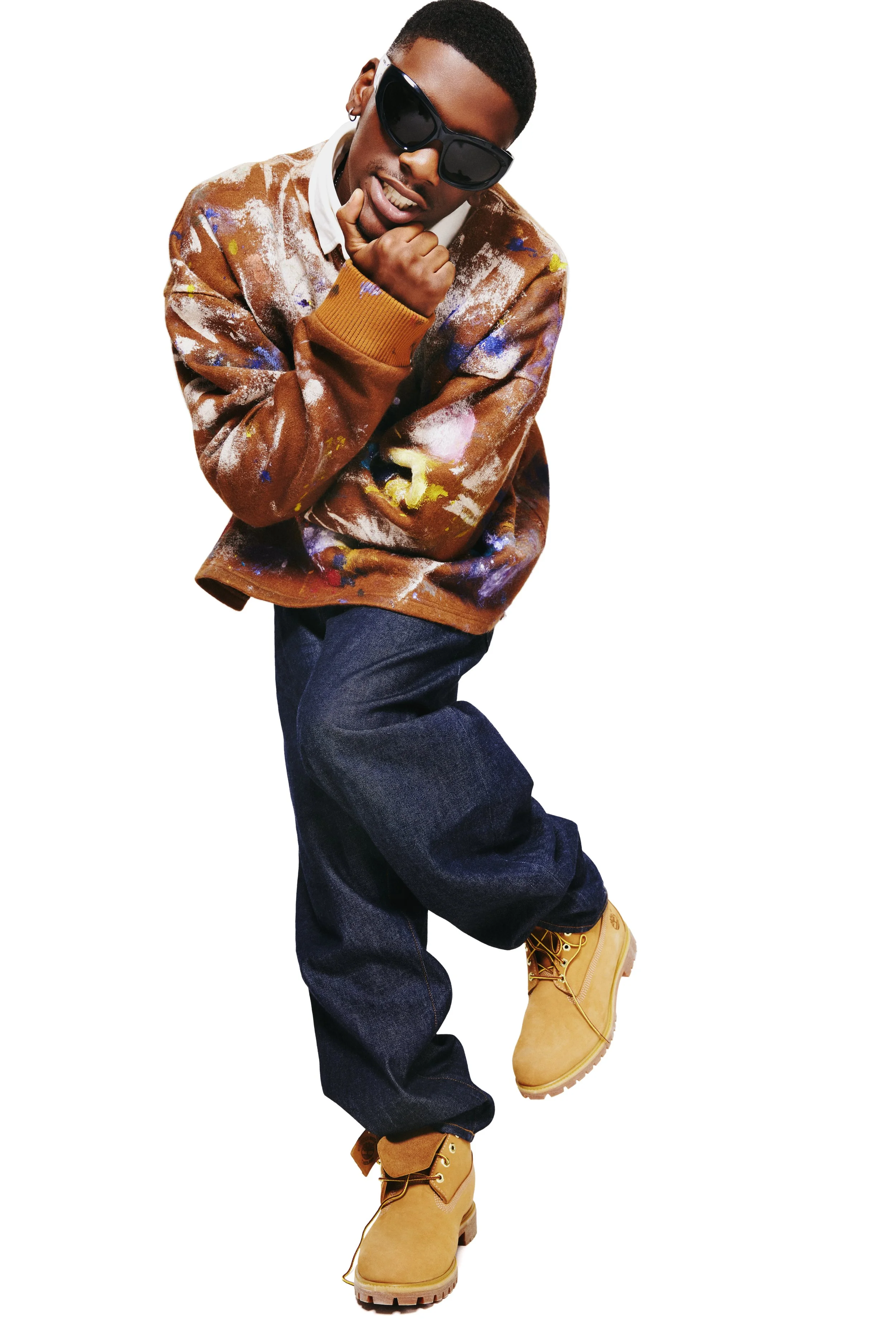 A man wearing black sunglasses, a multicolored paint-splattered jacket, dark jeans, and tan work boots, striking a playful pose against a white background.
