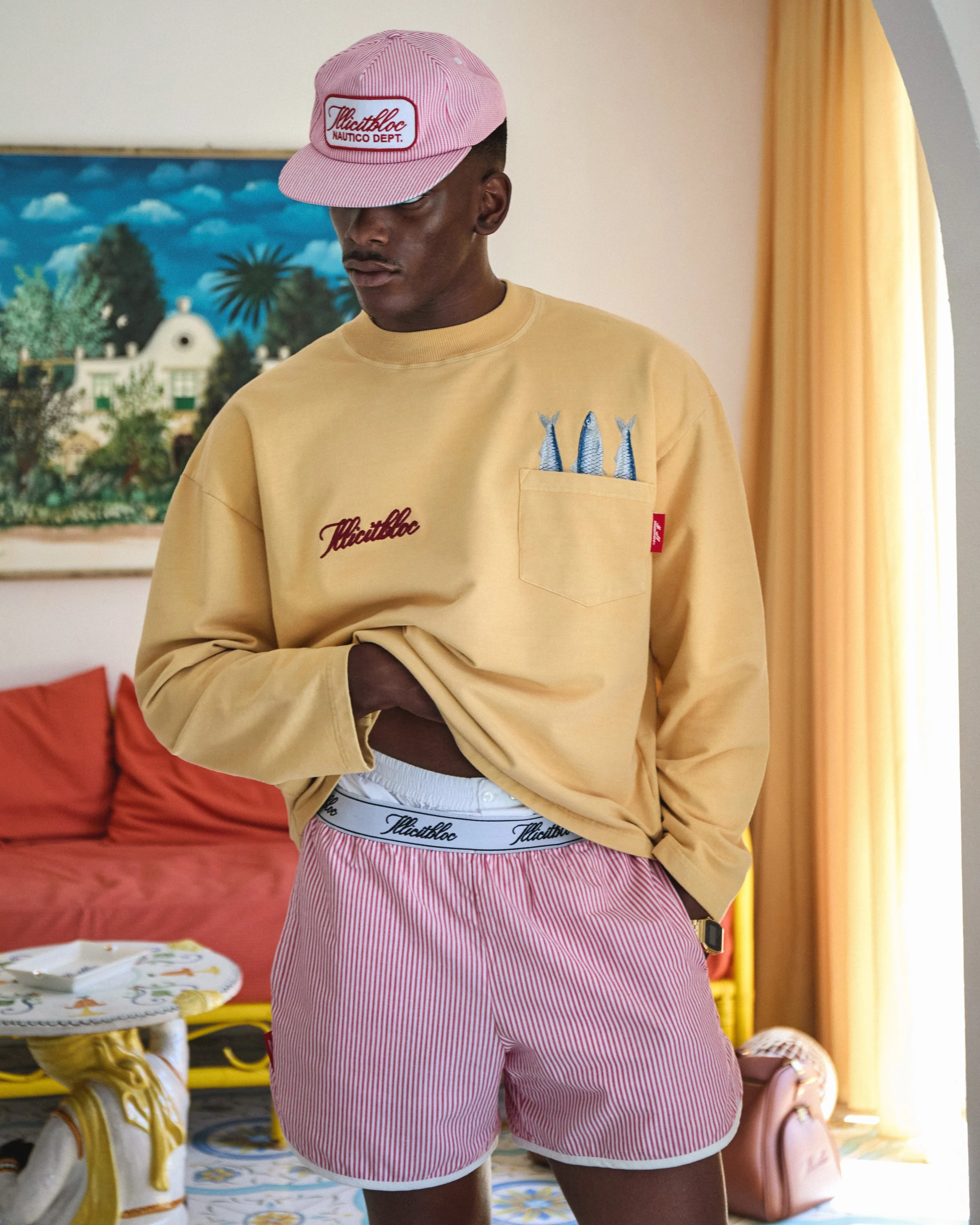 A man wearing a pink cap, yellow sweatshirt with embroidered fish and text, pink and white striped shorts, standing indoors with artwork and curtain in the background.
