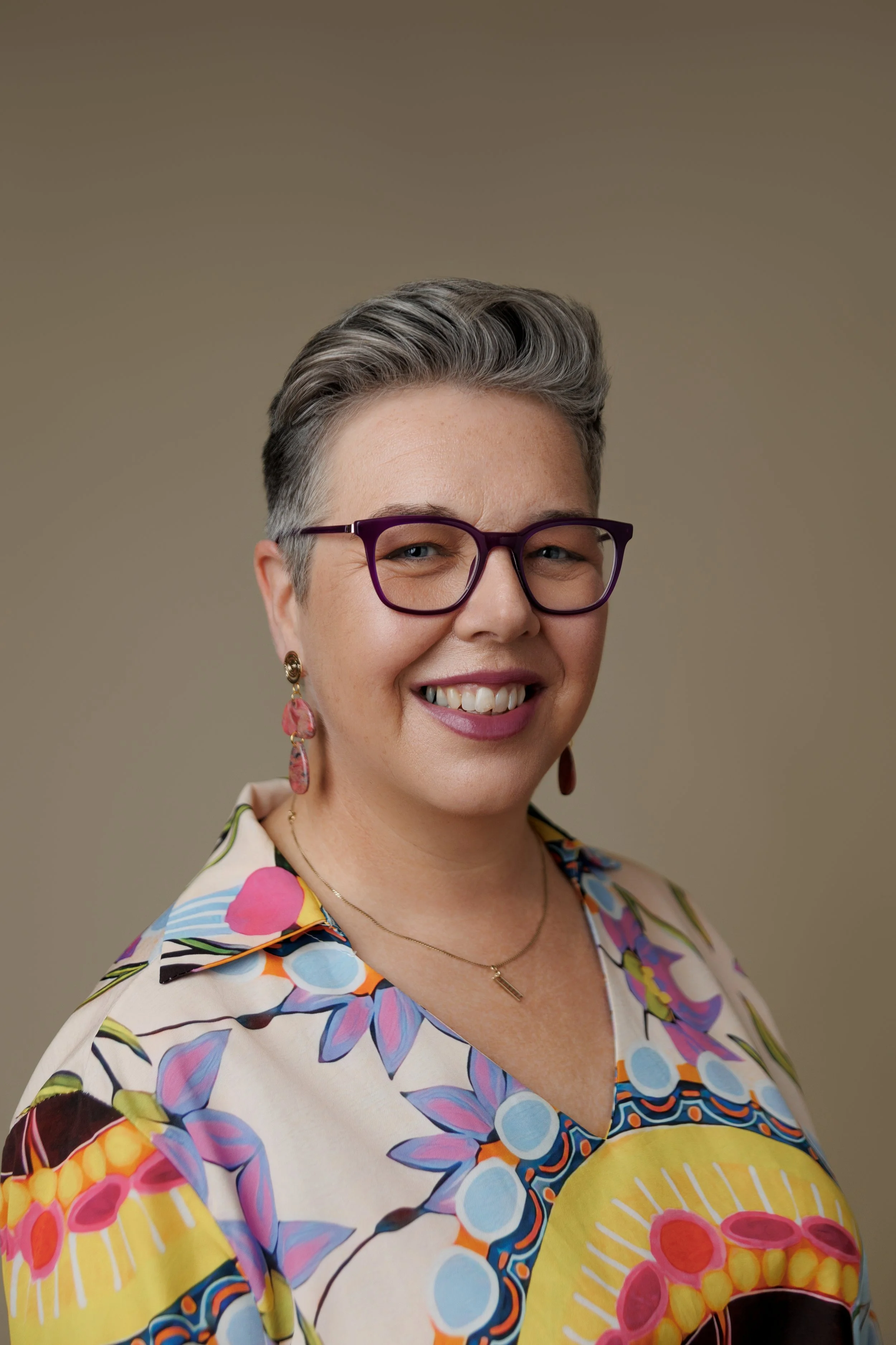 A woman with short gray hair, wearing purple glasses, colorful floral shirt, and pink earrings, smiling against a neutral background.