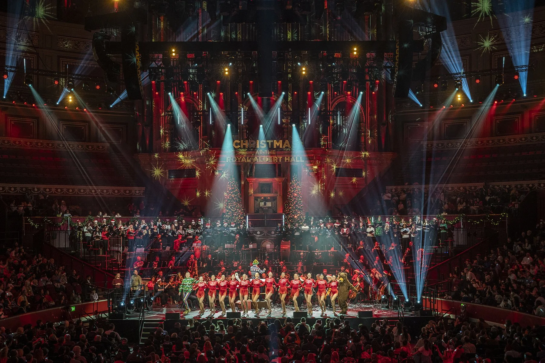 A stage decorated for Christmas with a choir, performers, and dancers celebrating during a Christmas show with colorful lighting and audience in attendance.