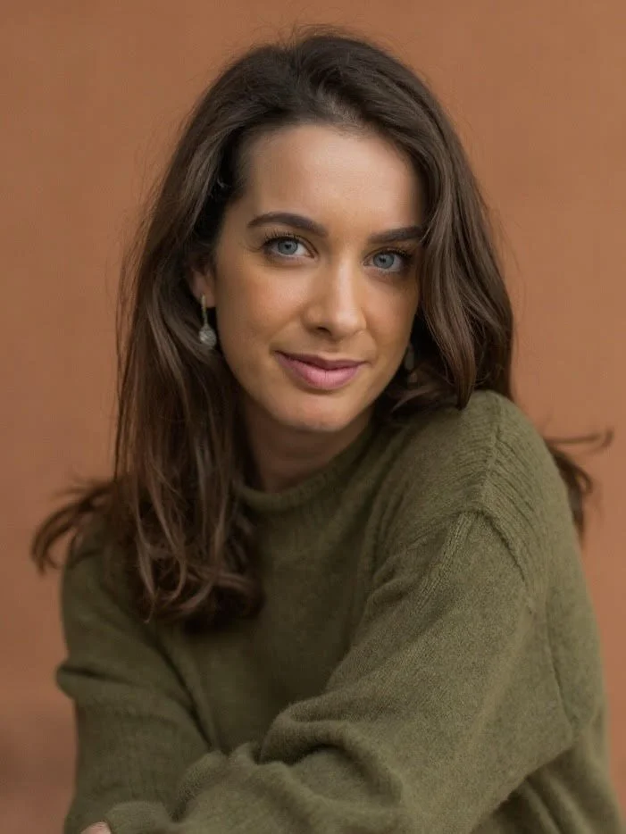 A young woman with long brown hair and blue eyes, wearing a green sweater and earrings, smiling softly with a warm background.