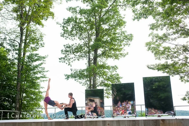 A performer doing a handstand while a person holds their legs during an outdoor show on a wooden platform, with trees and three reflective panels behind them.