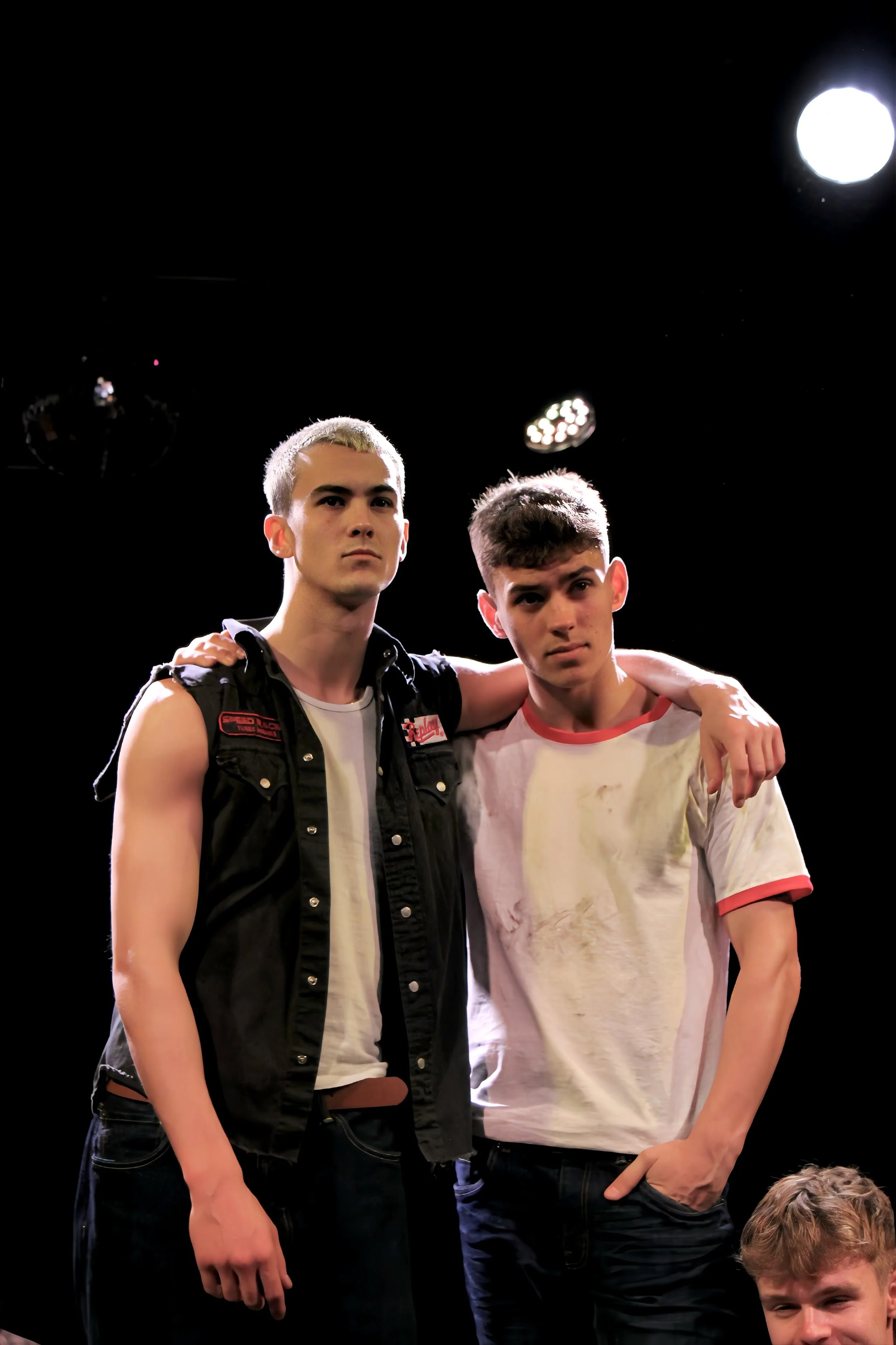Two young men standing on stage with a black background and bright stage lights, one with arm around the other's shoulder.
