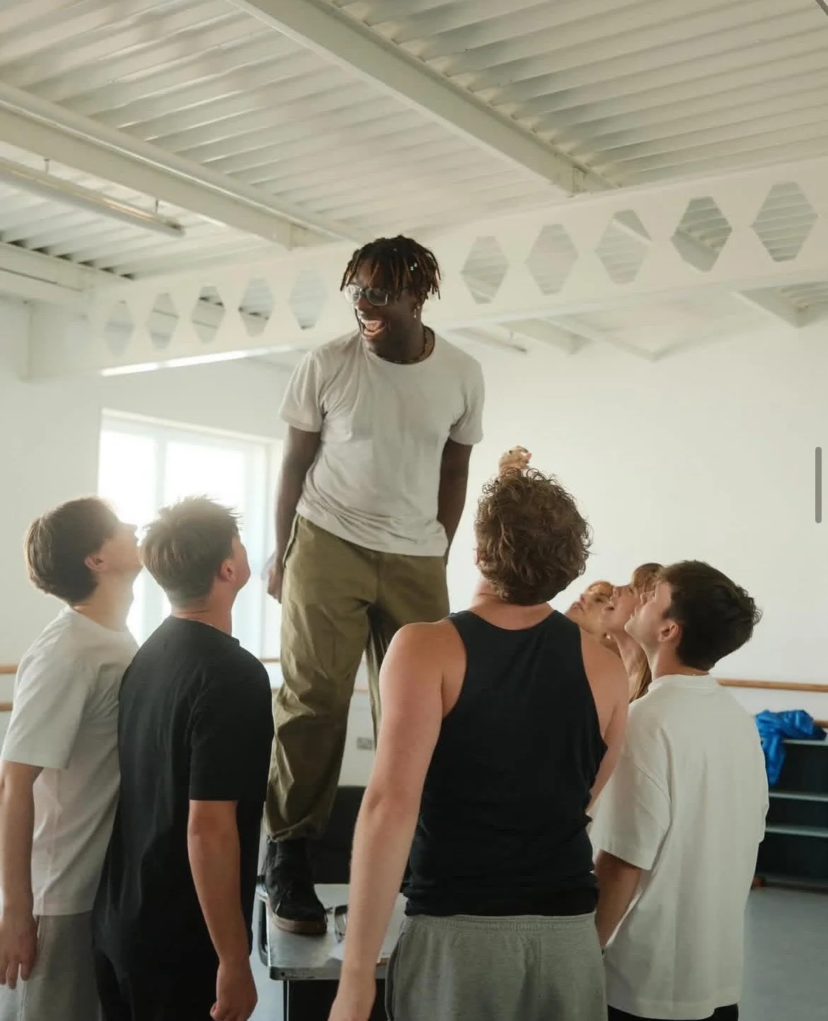 Group of people engaging in a team-building activity, with one person standing on a table and others supporting him, in a bright room.