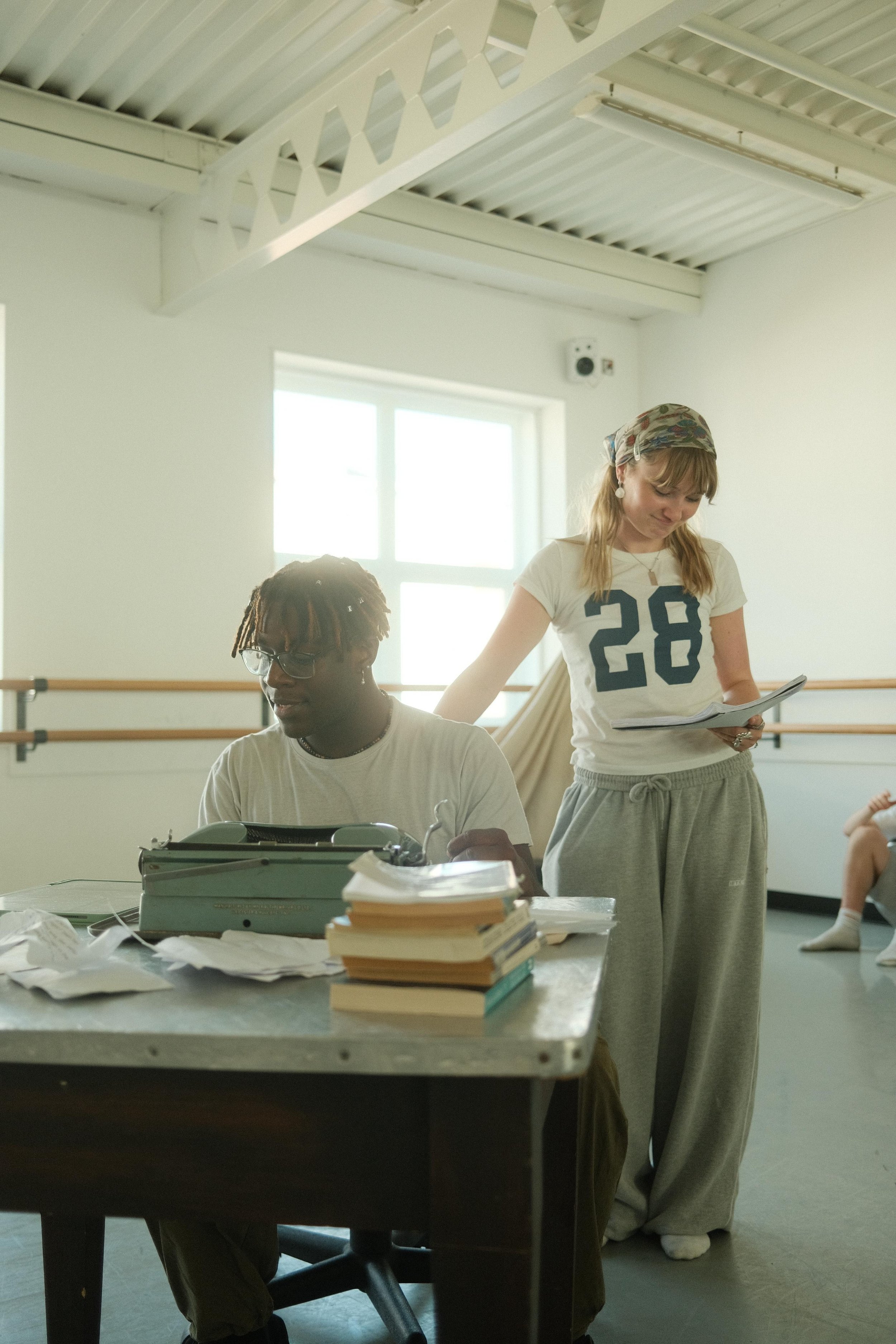 Two women working at a desk with papers and a typewriter, one sitting and one standing, in a bright room with large windows and ballet barres.