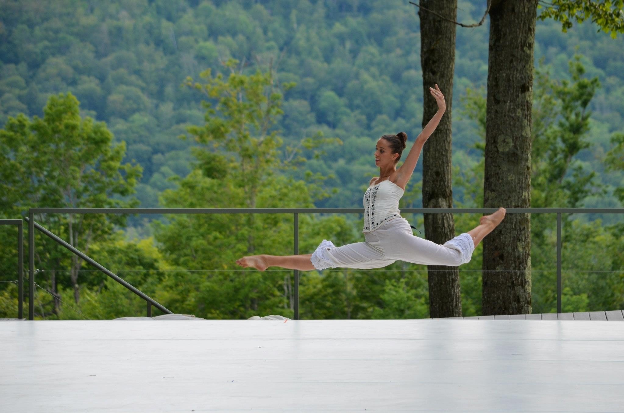 A woman in white pants and a white top performs a ballet leap outdoors on a stage with a green, forested background.