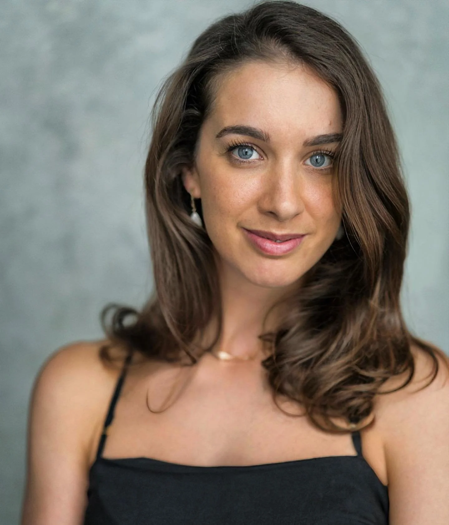 A portrait of a woman with blue eyes, wavy brown hair, wearing a black spaghetti strap top, and a delicate necklace, smiling softly against a neutral background.