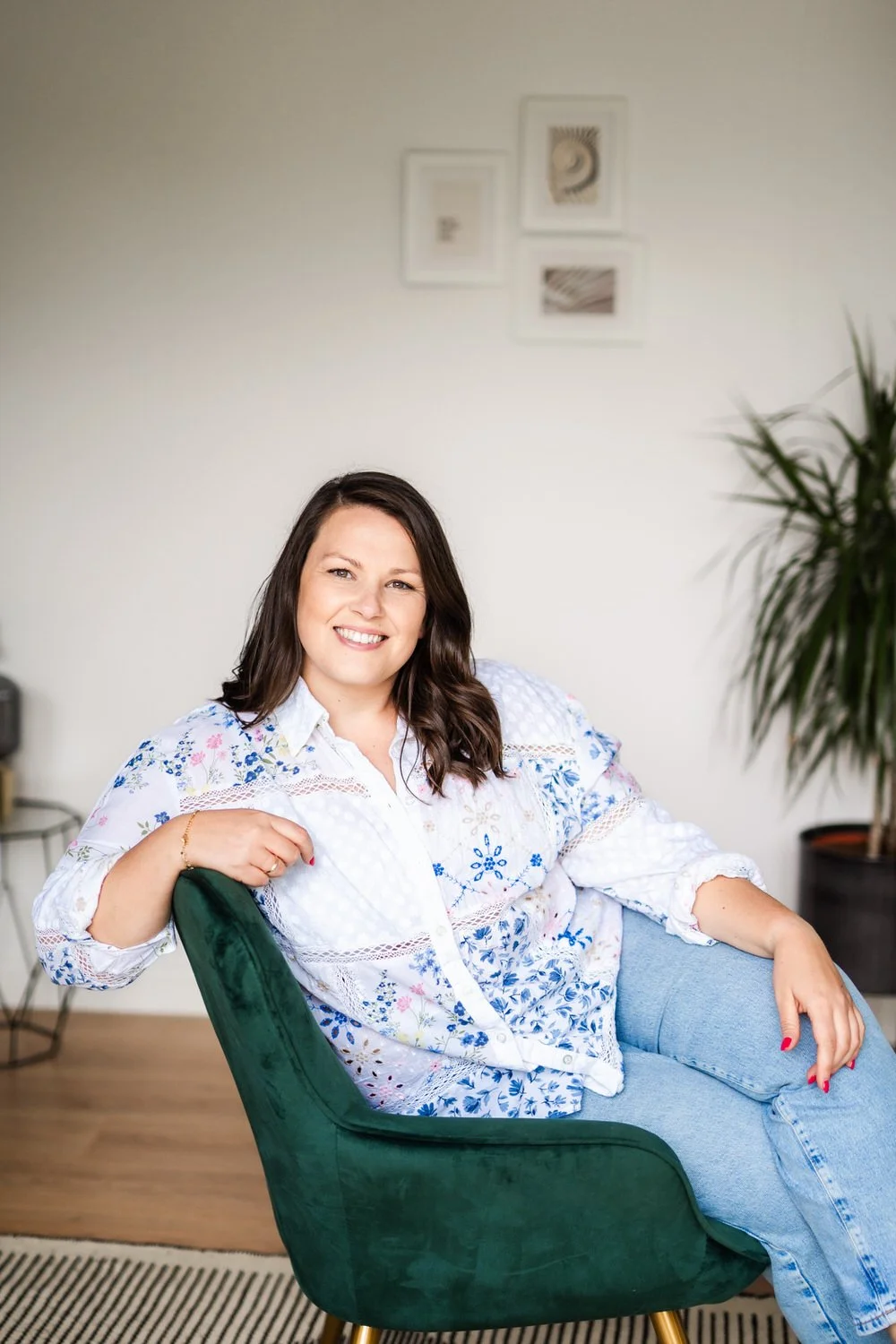 Smiling woman with brown hair sitting on a green chair in a bright room with a white wall and framed artwork, a potted plant in the background.