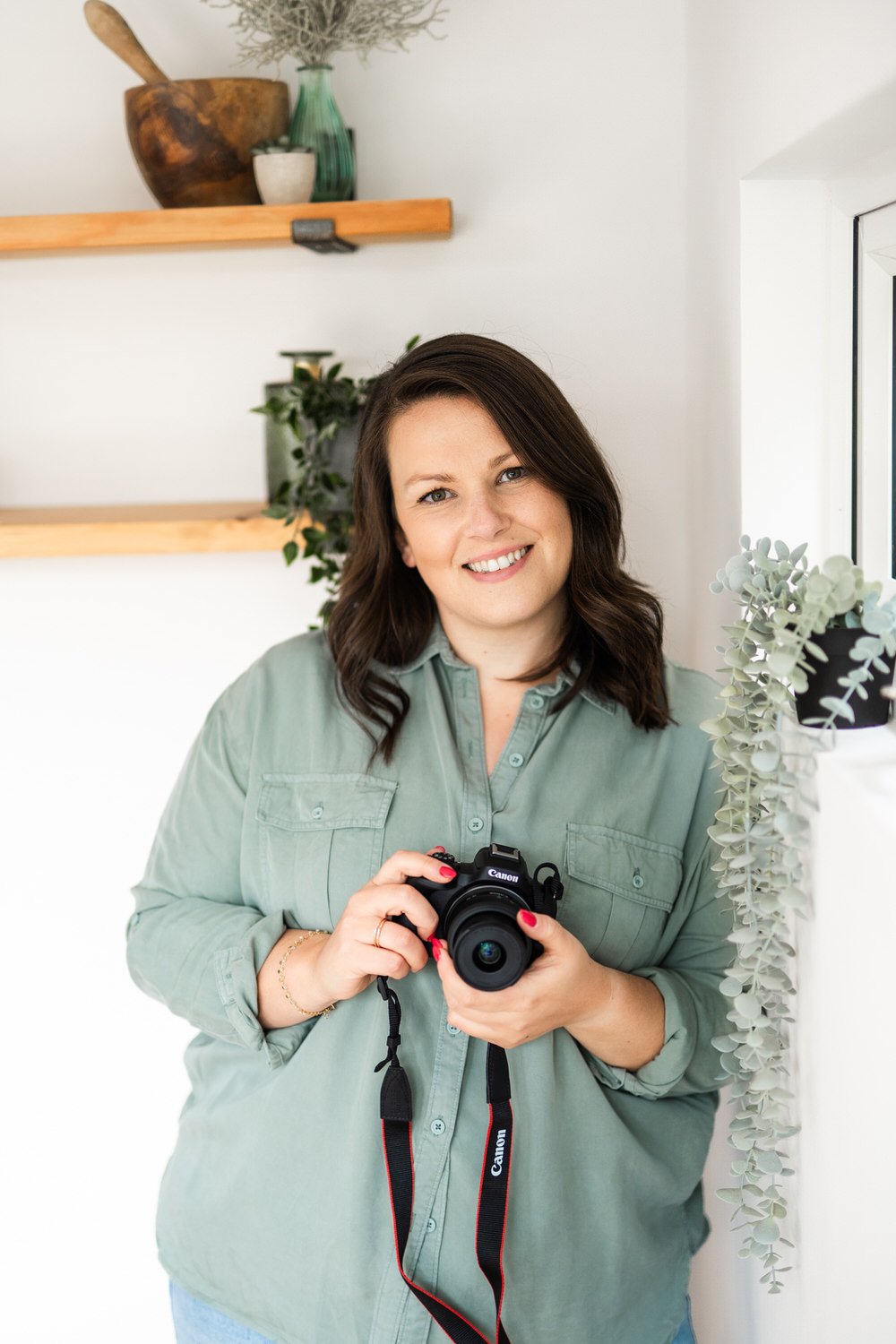 A woman with dark brown hair, smiling, holding a black Canon camera, standing indoors near a mirror with a white and green decor background.
