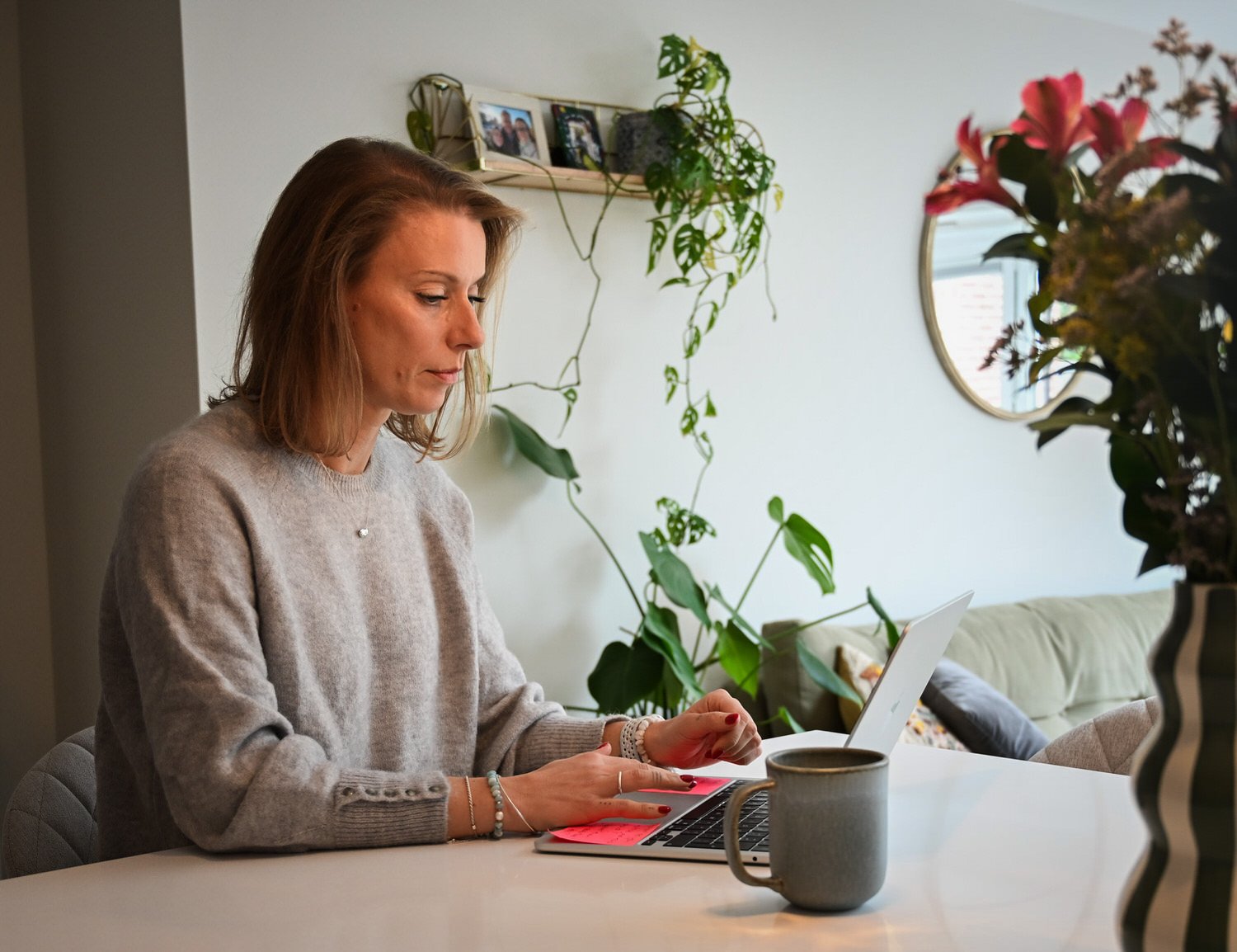 A woman sitting at a kitchen table using a laptop with a red cover, a mug nearby, and houseplants in the background.