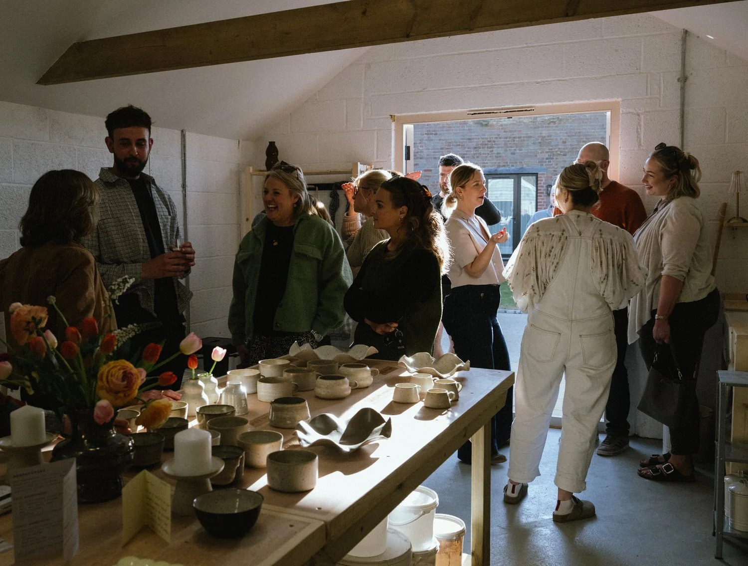 Group of people gathered inside a room with sunlight coming through the door opening, engaging in conversation. Table with pottery and flowers in the foreground.