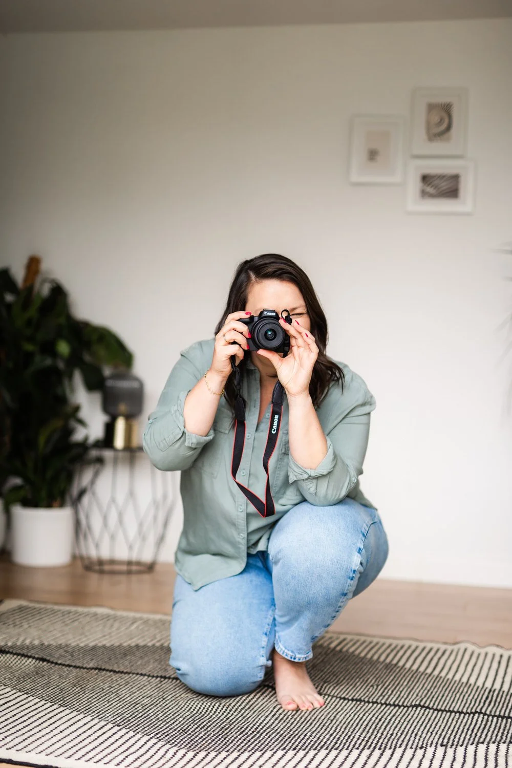 A woman kneeling on a rug, taking a photo with a DSLR camera inside a home.