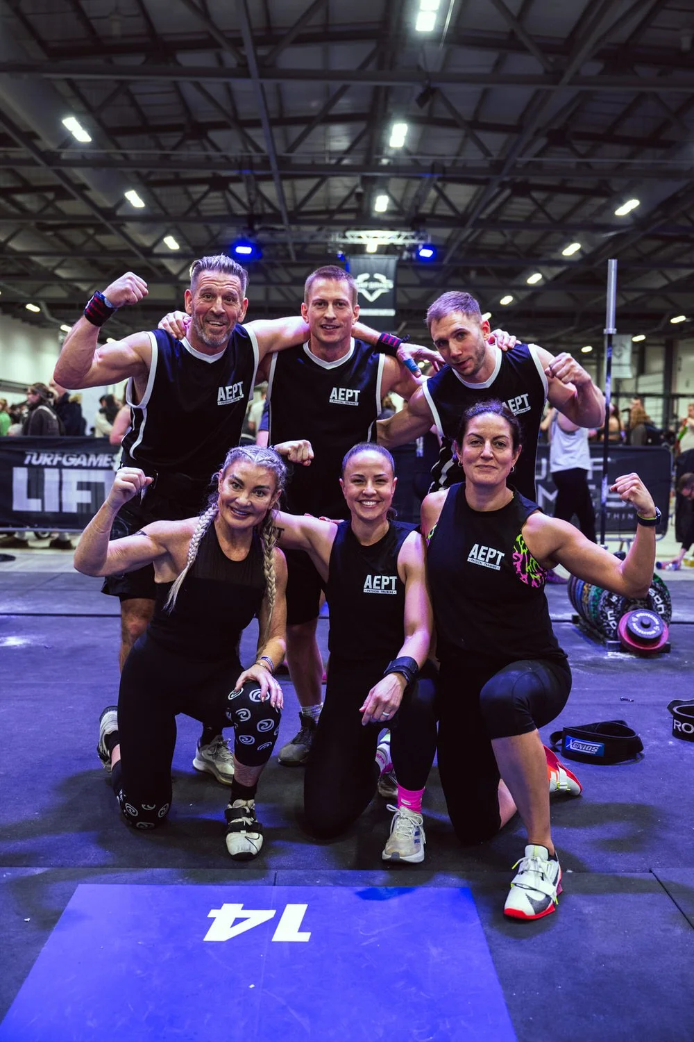 Group of six fit people, three men and three women, in athletic gear posing for a photo at a fitness event. They are flexing their arms and smiling, with a gym or fitness competition setting in the background.