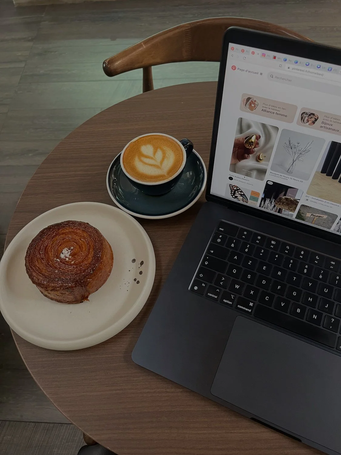 A coffee cup with latte art, a croissant on a white plate, a laptop, and a cup of coffee are on a round wooden table in a cafe.
