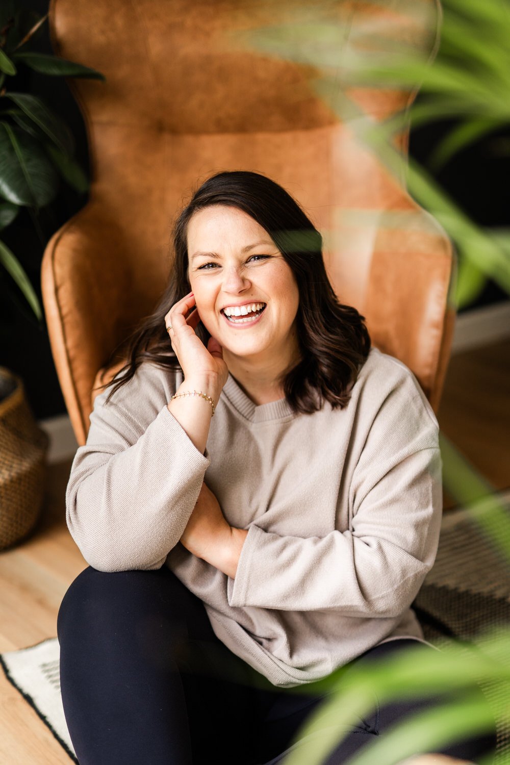 A woman with dark brown hair and light skin is sitting on the floor, leaning against a tall, brown leather armchair, smiling and laughing while touching her ear. She is wearing a beige sweatshirt and black pants, with a plant partially visible in the foreground and background.